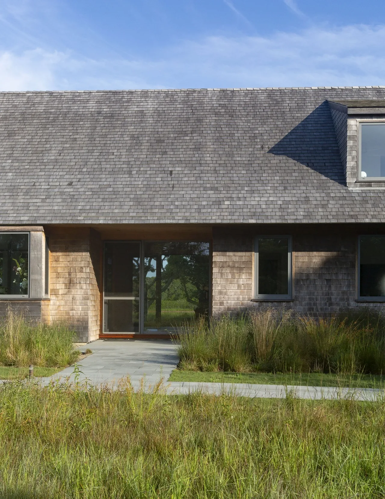 The Wink House features wood shingle siding, a dormer window, and a stone walkway through natural landscaping with tall grasses.