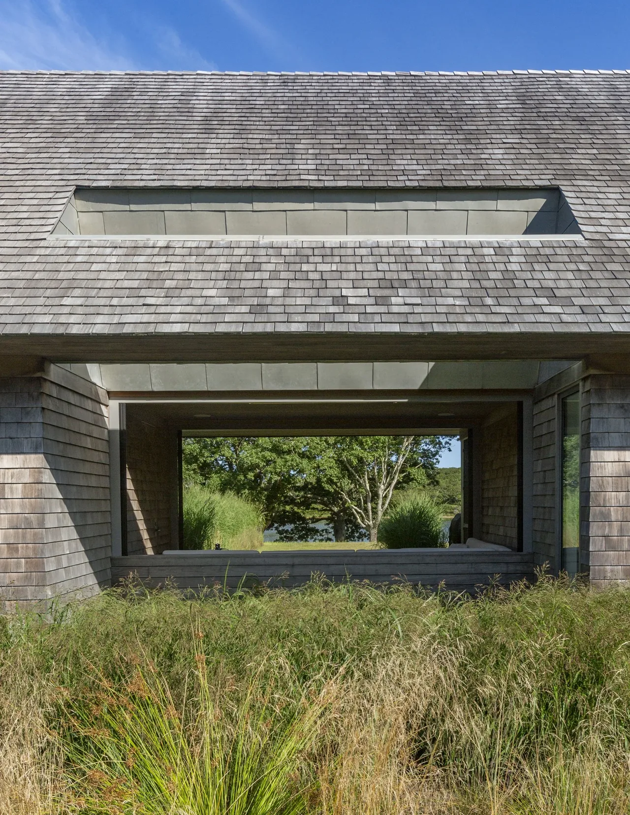 Wink House features a pitched roof with cedar shingle siding and a large horizontal window opening framing natural landscaping.
