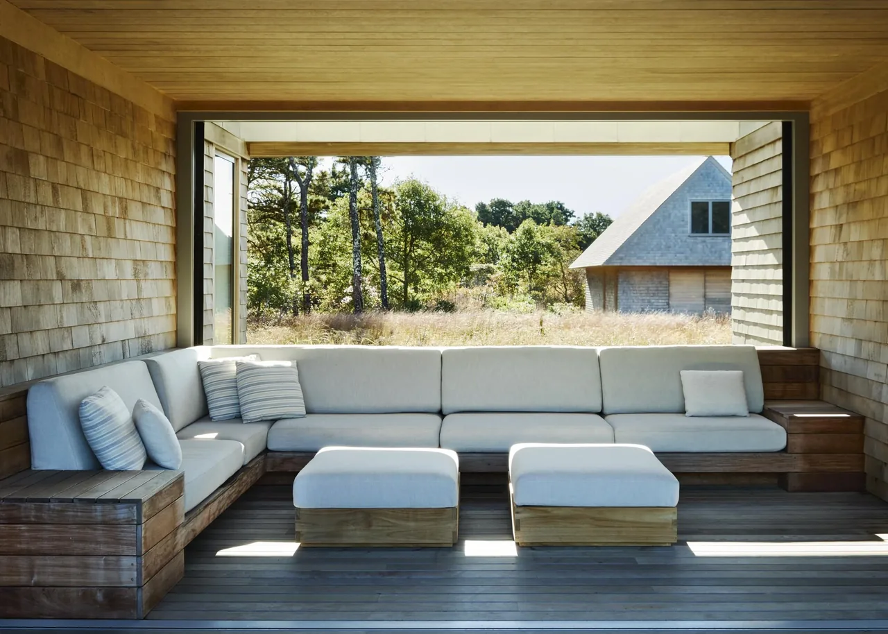 The Wink House features a covered outdoor living area with built-in wooden seating and a large horizontal window opening to the natural landscape.
