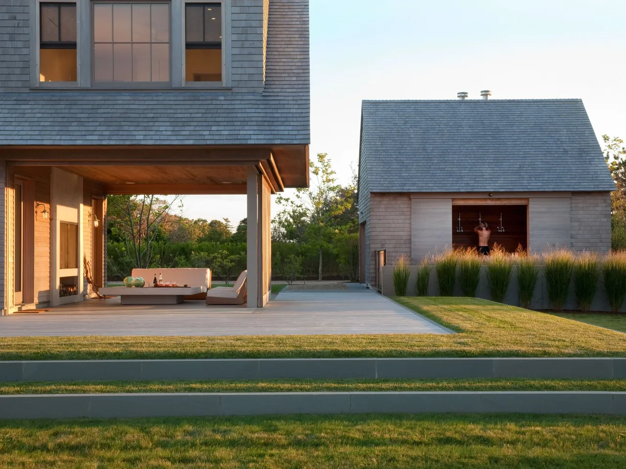 Patio seating under a covered porch next to a small building with an open shower in a landscaped yard during sunset.