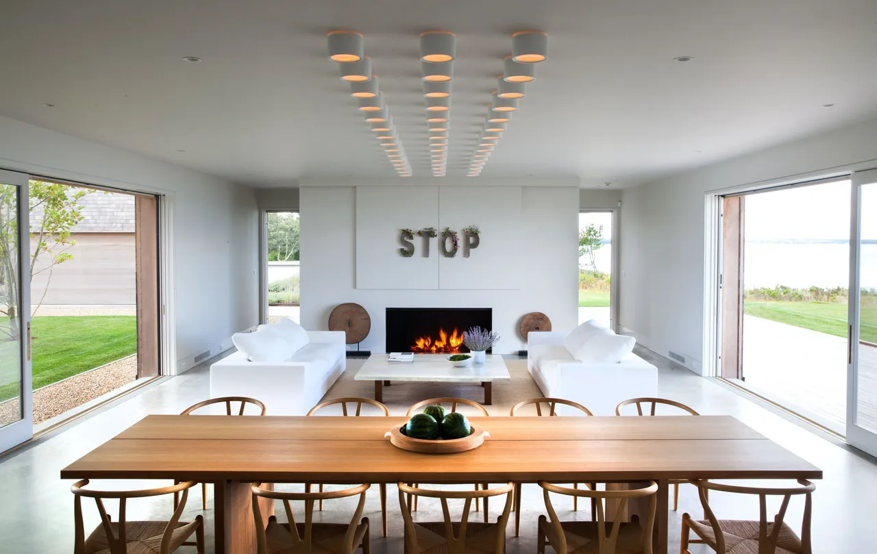 Open living and dining space with fireplace, white seating, wood furniture, and ocean view through wide glass doors in Island House by Noury-Ello Architects.