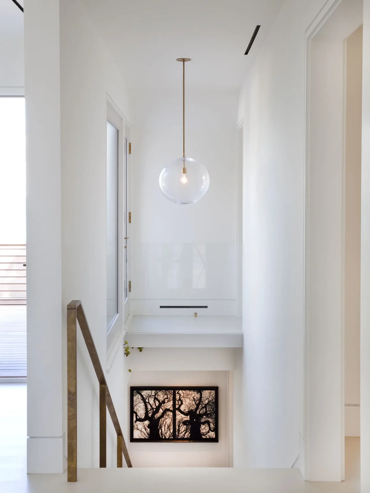 Minimalist white-walled staircase landing features a clear globe pendant light and silhouetted tree artwork below.