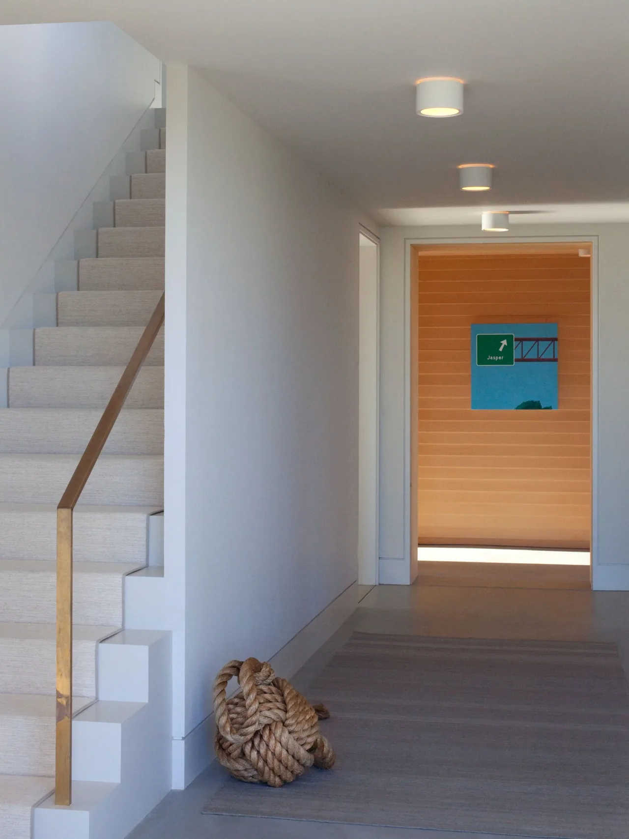 Hallway view shows beige carpet stairs with brass handrail and natural fiber rug with coiled rope at foot of stairs