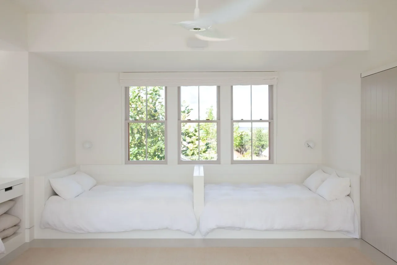Island House bedroom by Noury-Ello Architects with twin beds and white walls featuring three flanked windows.