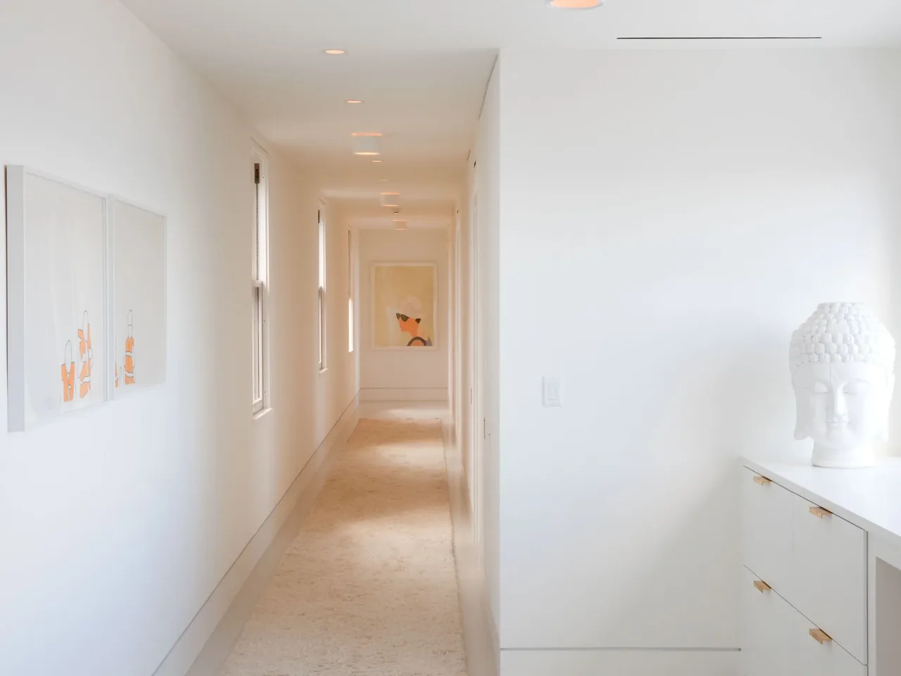 Minimalist white hallway features recessed lights and framed artwork with a large decorative sculpture on a cabinet.