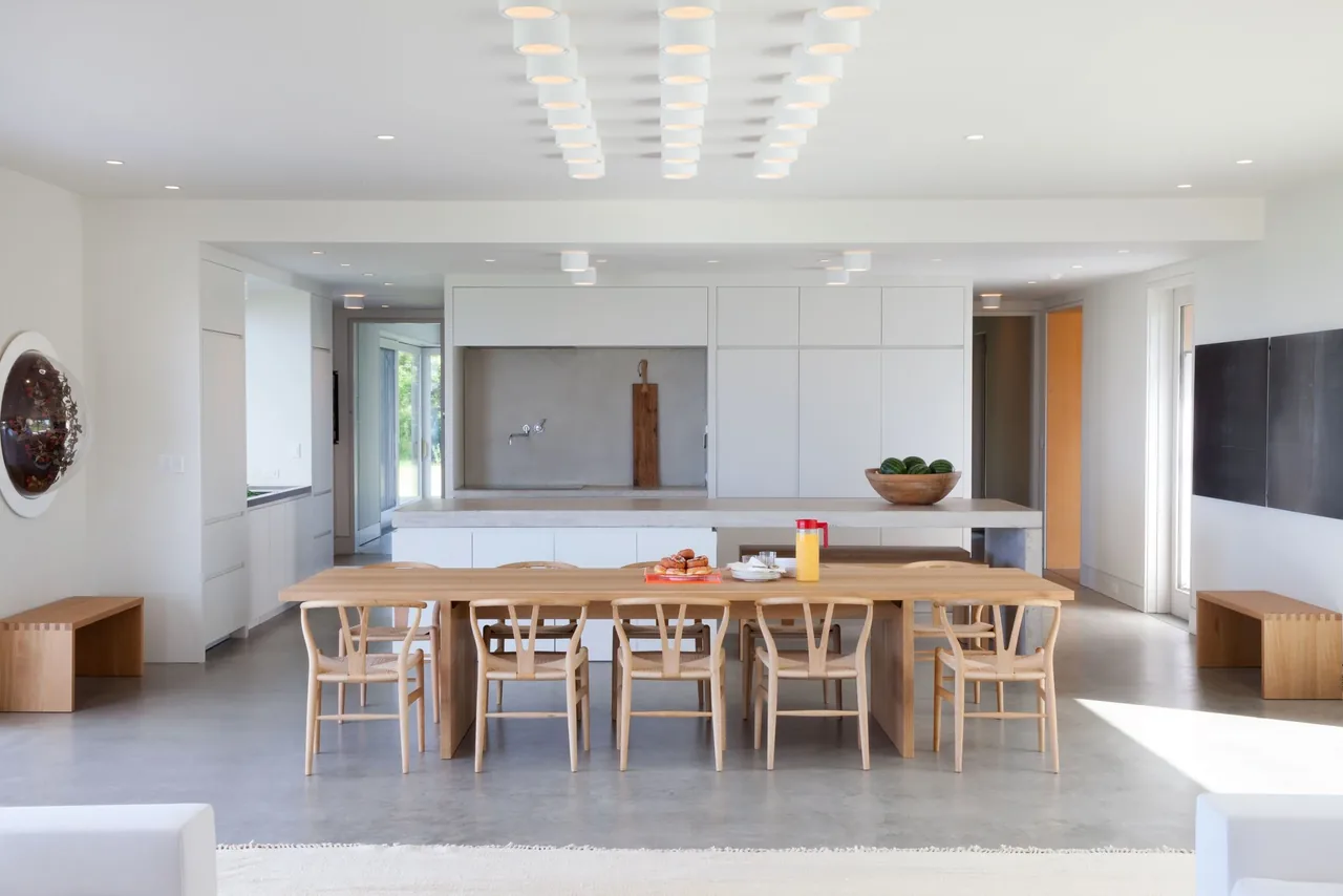 The Island House kitchen and dining area features a long wood table, white cabinetry, and minimalistic ceiling lighting.