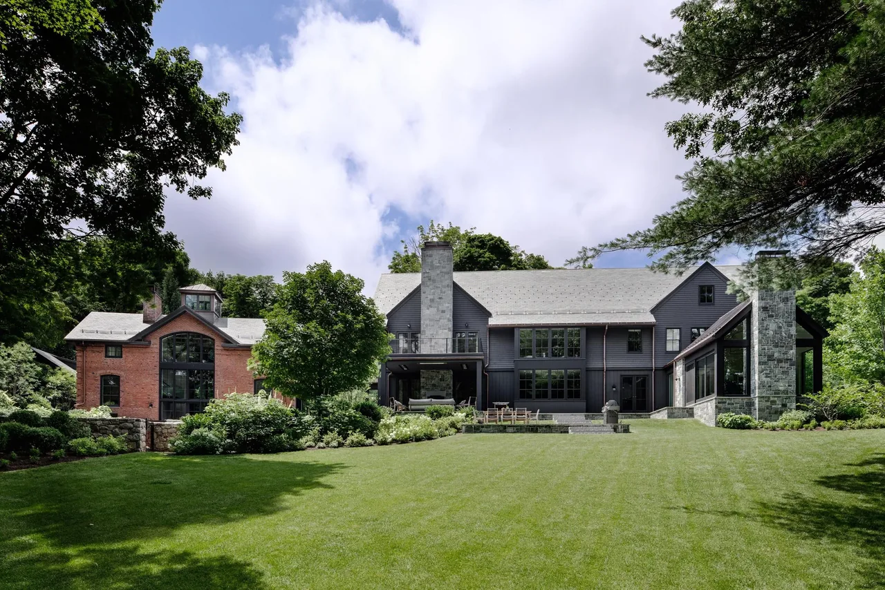 Black-sided main house with large windows and stone chimneys connected to a brick carriage house with arched window.