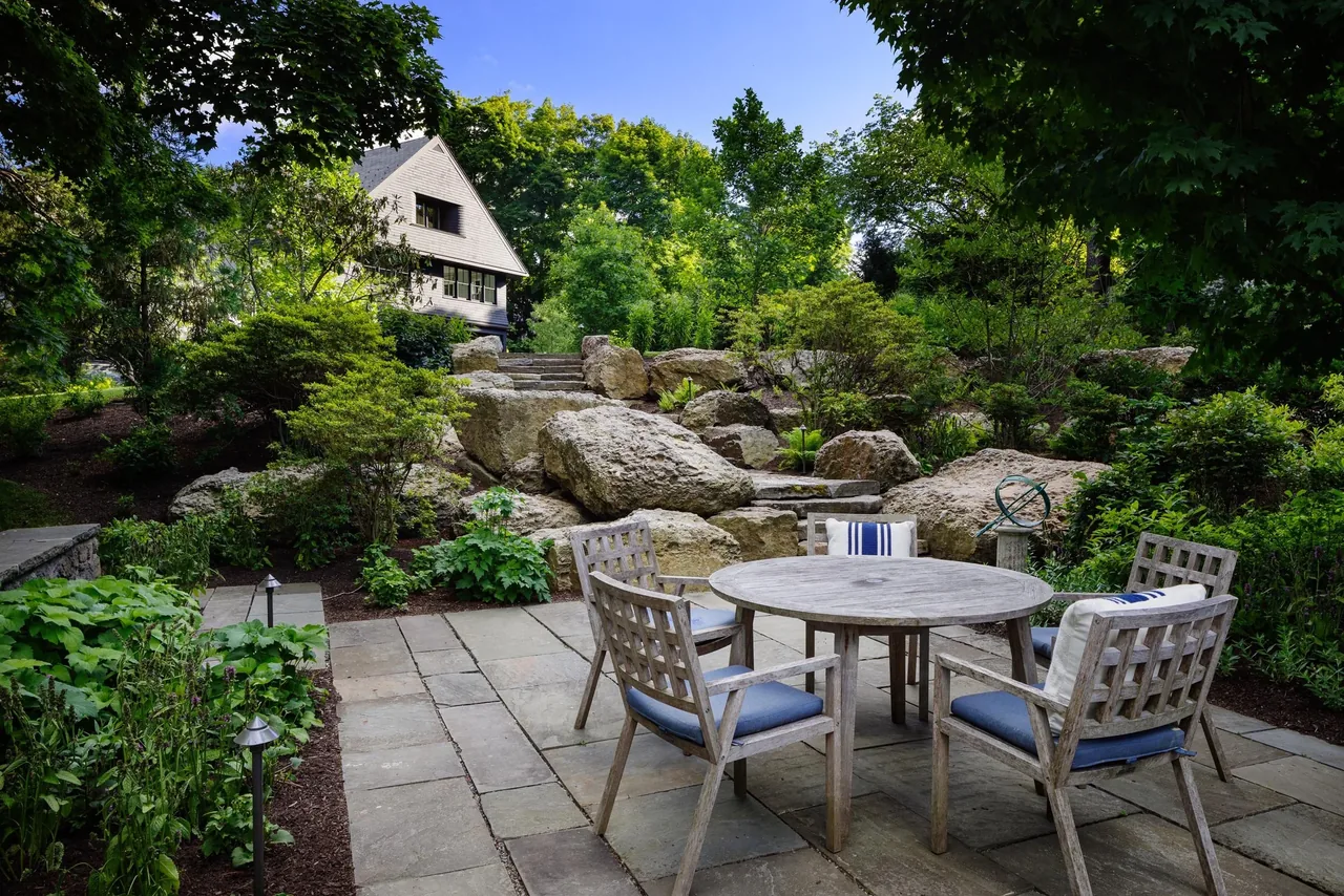 Stone patio with wooden outdoor furniture set amidst a lush, landscaped garden and large rocks.