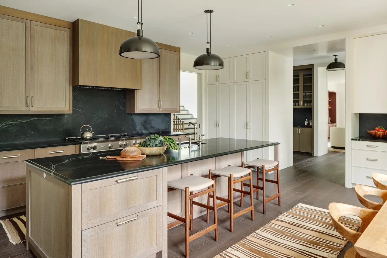 Modern kitchen features light wood and white cabinetry with a black stone island countertop and pendant lighting.