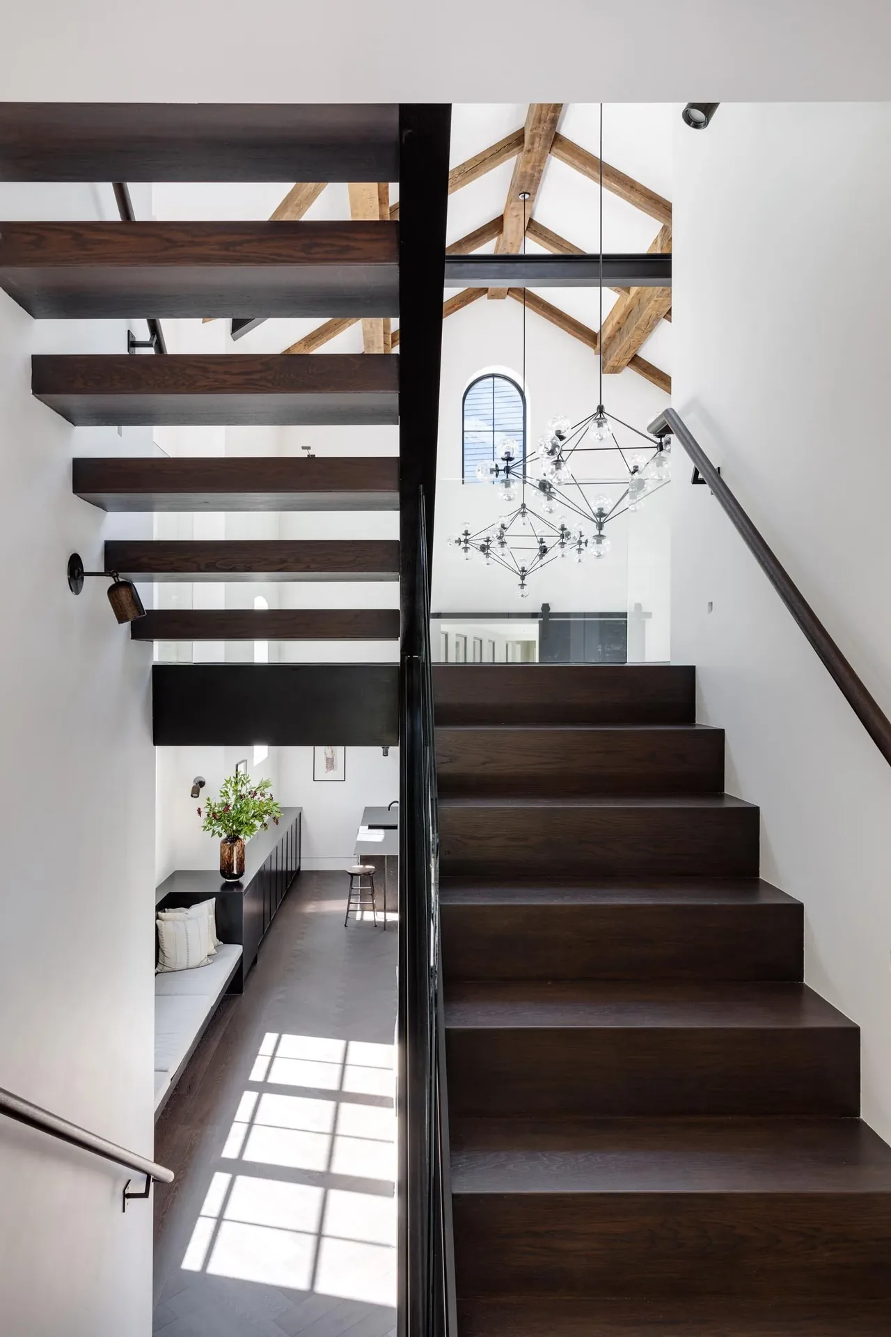 Dark wood staircase with glass balustrade and black metal chandeliers beneath exposed wood roof beams.