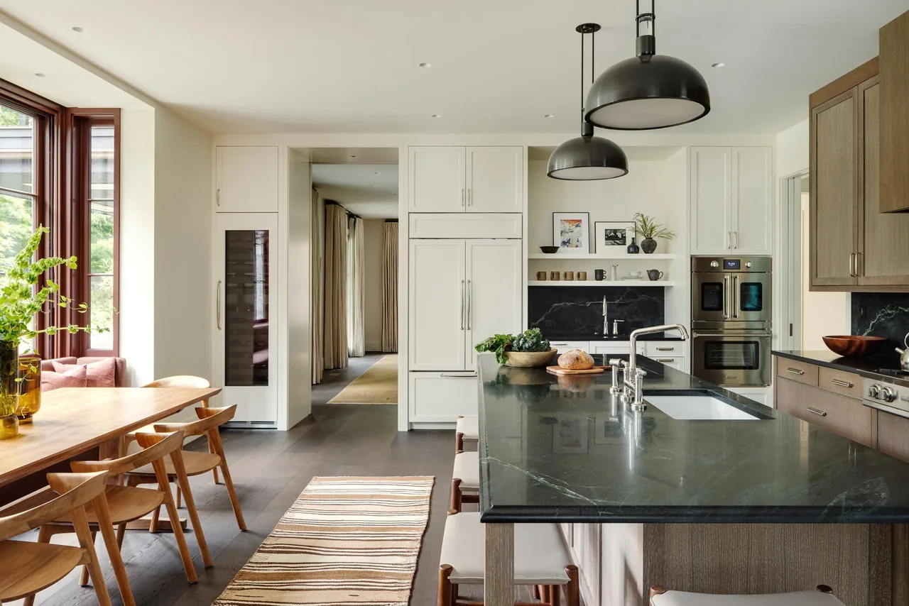 Kitchen features a large black stone island with seating, white cabinetry, built-in appliances, and a wooden dining table by a window.
