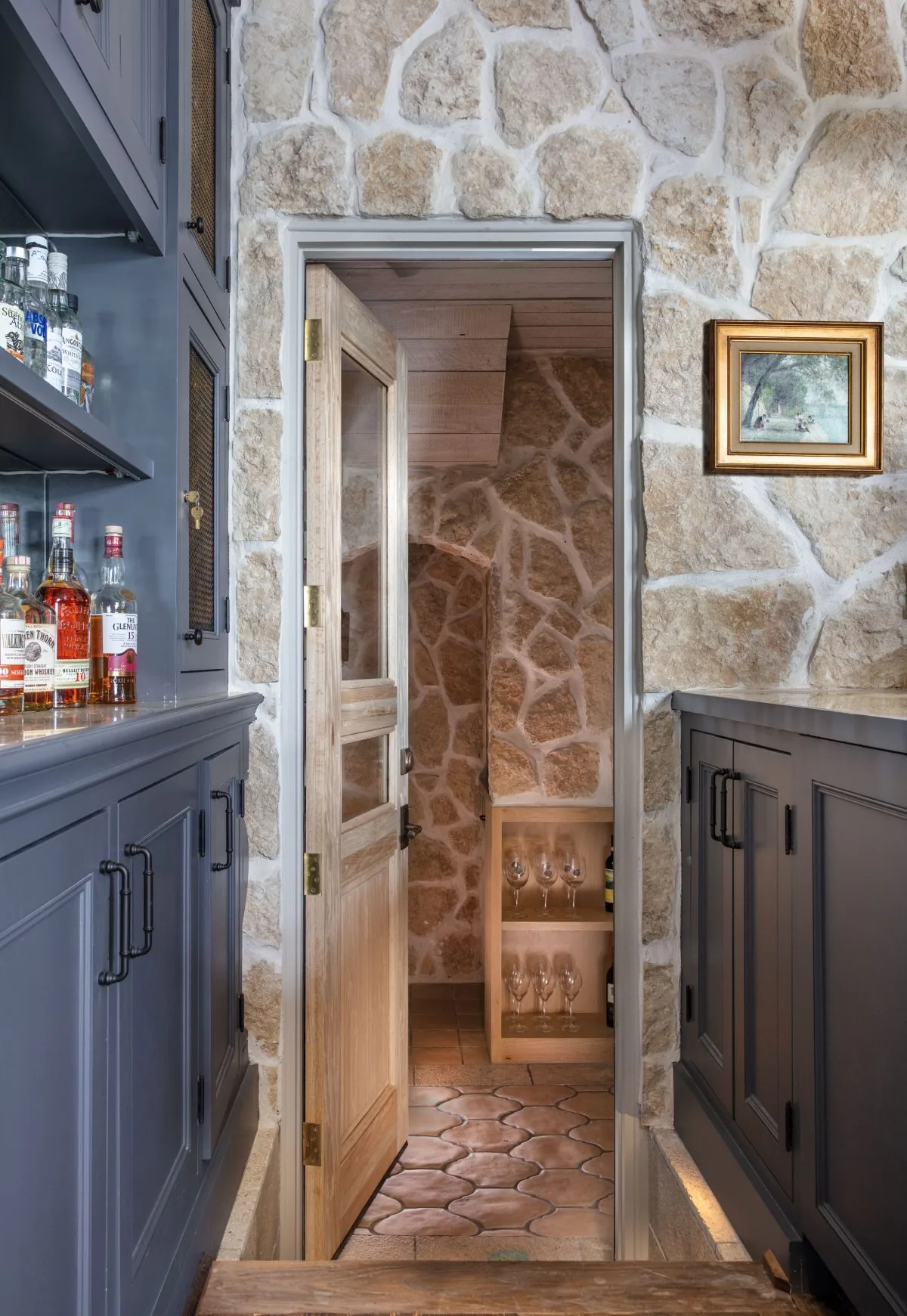 A home bar featuring stone walls, a glass door, and built-in cabinetry leads to a wine storage room with terracotta tile flooring.