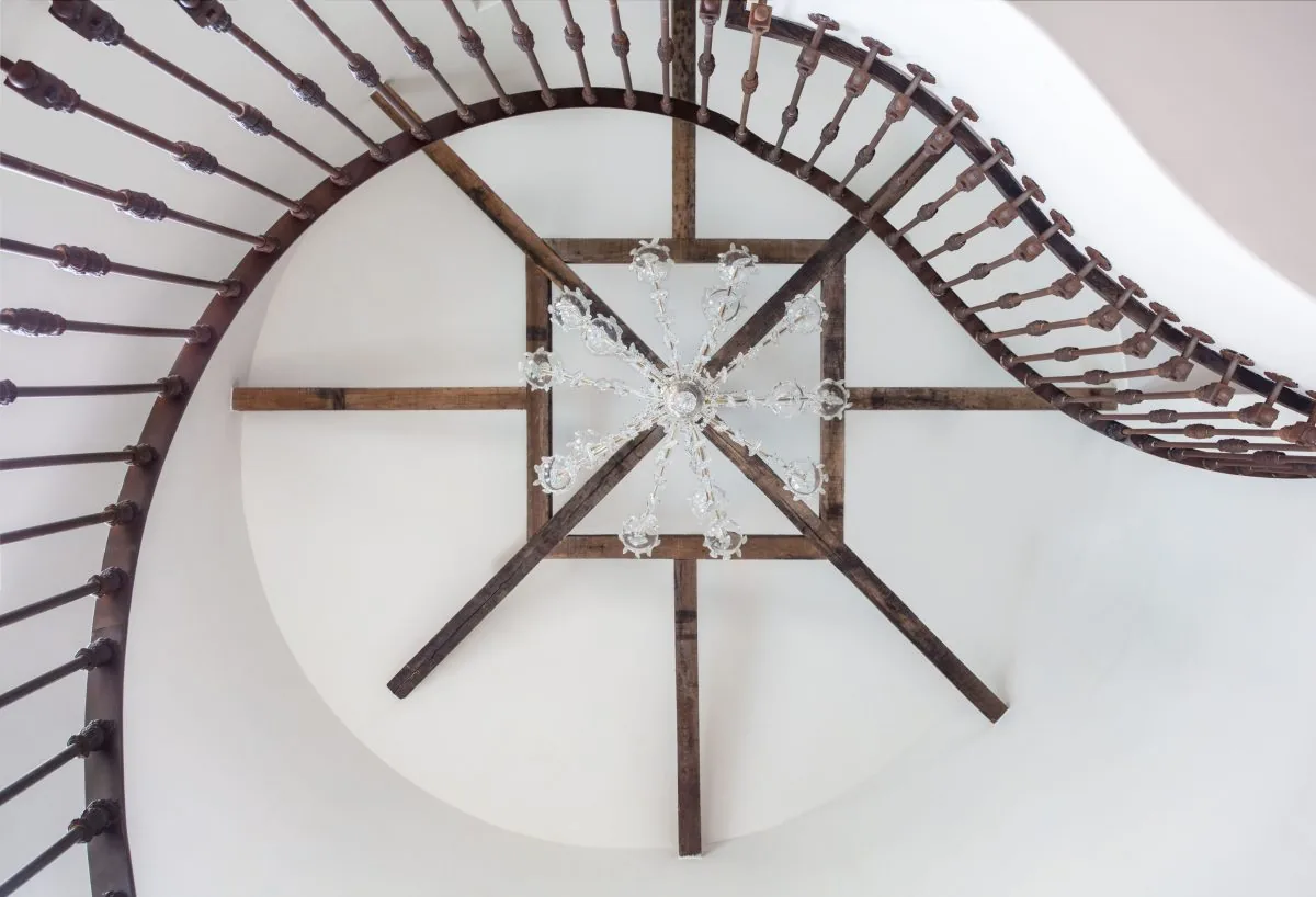 Curved staircase with wooden balusters and a chandelier centered on wood beams on the ceiling.