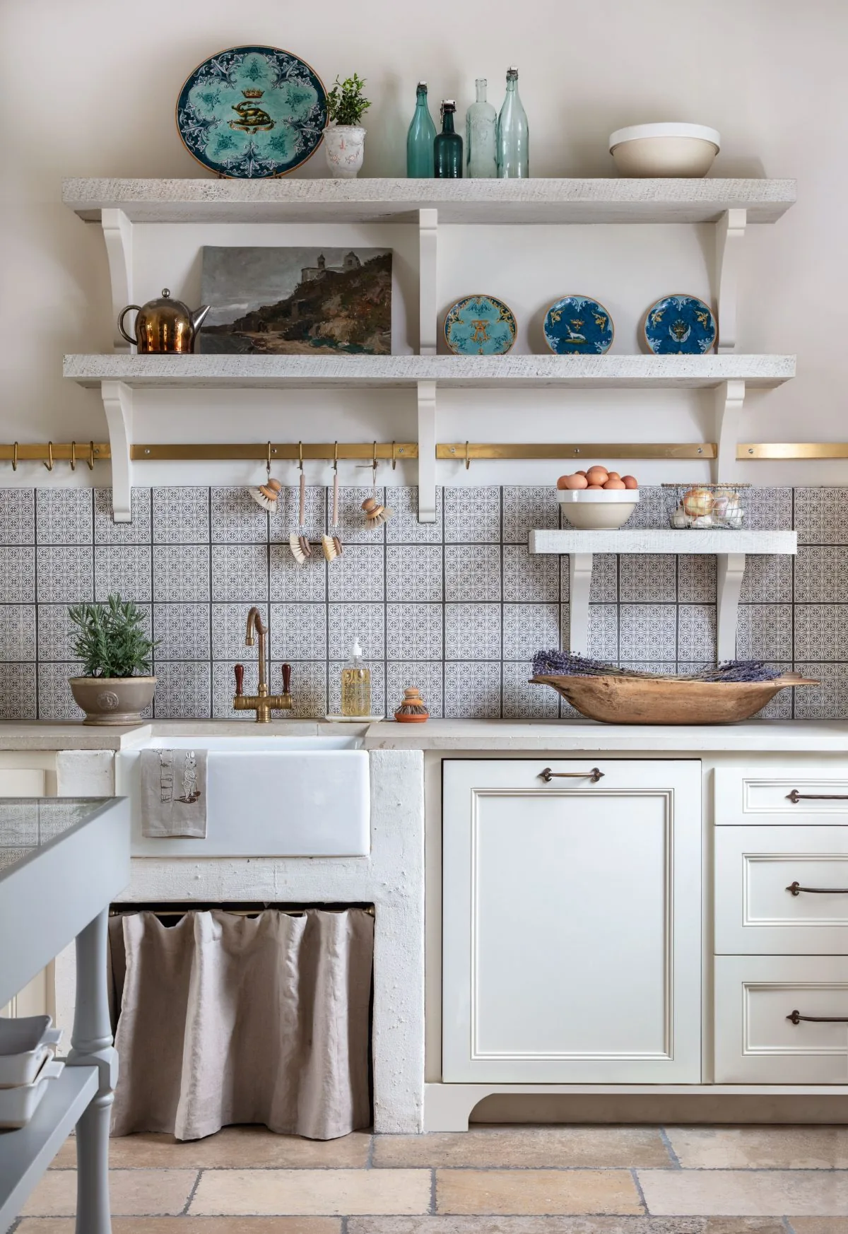Kitchen sink wall with open shelving displaying plates and bottles, featuring a farmhouse sink and brass faucet.
