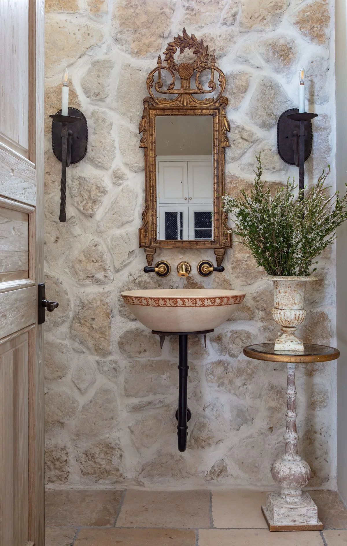 A Old-World French Estate powder bath features a stone wall, an ornate framed mirror, and a ceramic bowl sink with wall-mounted faucets.