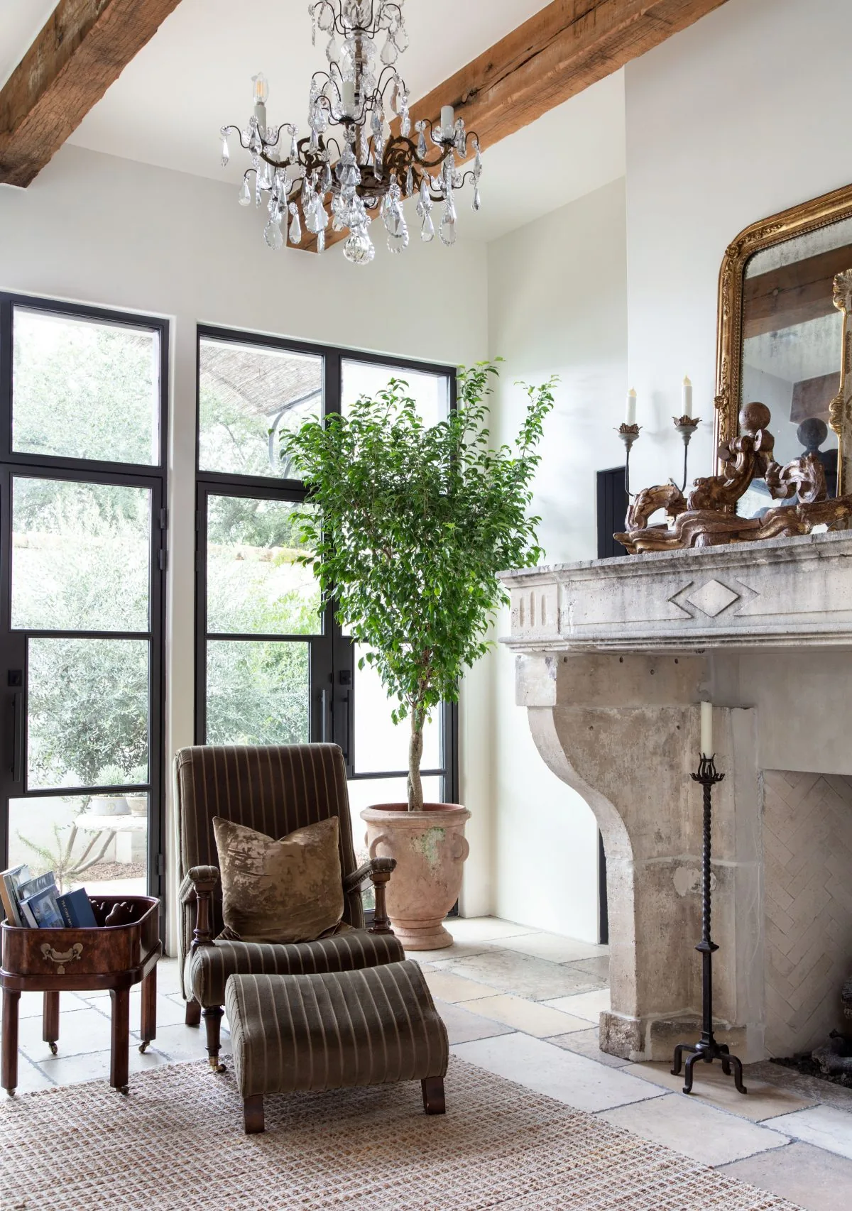 Living area with rustic stone fireplace, striped chair and ottoman, large potted plant, and black-framed windows.