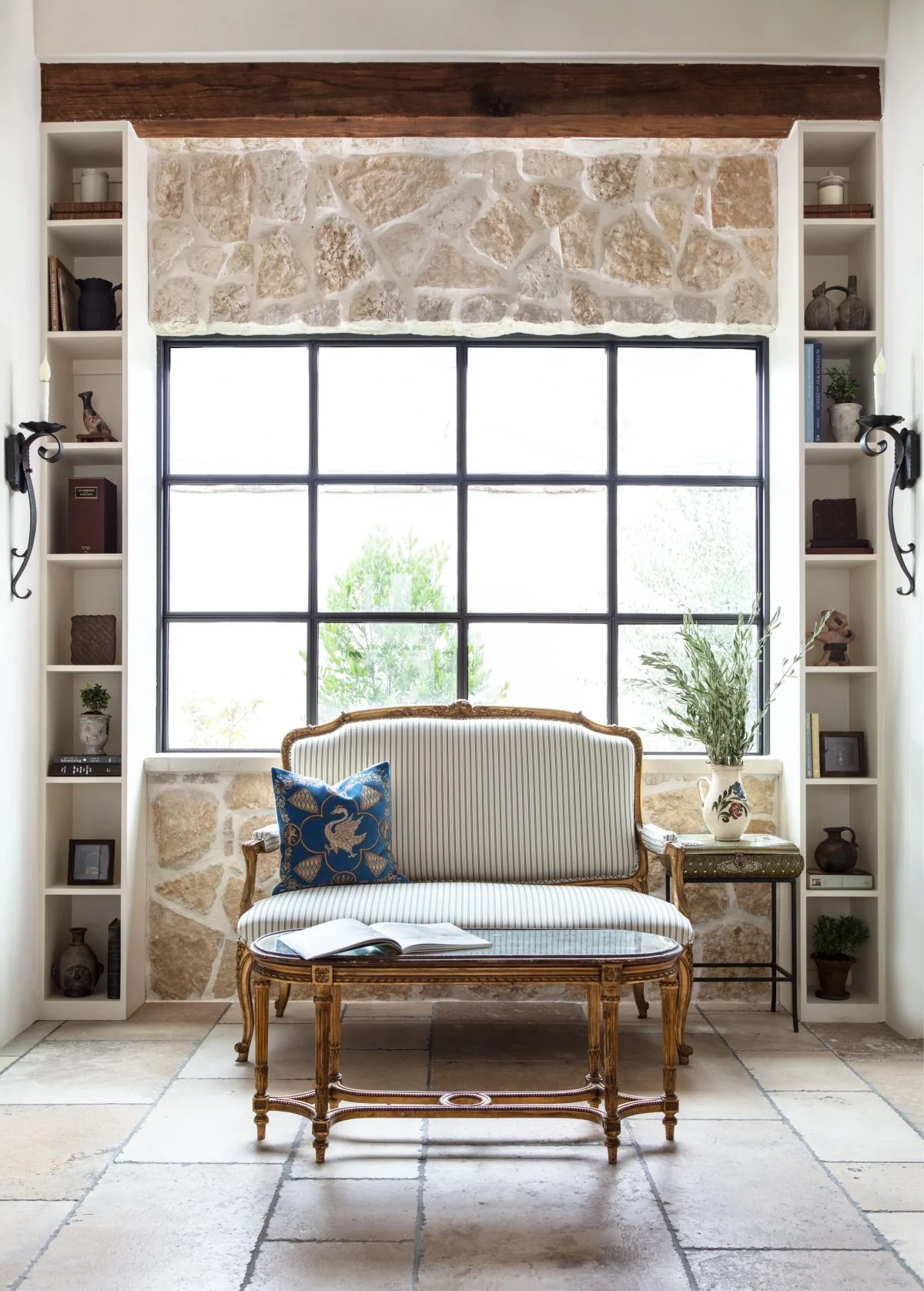 Sitting area with vintage-style settee and built-in shelving flanking a large black-framed grid window with stone wall surround.
