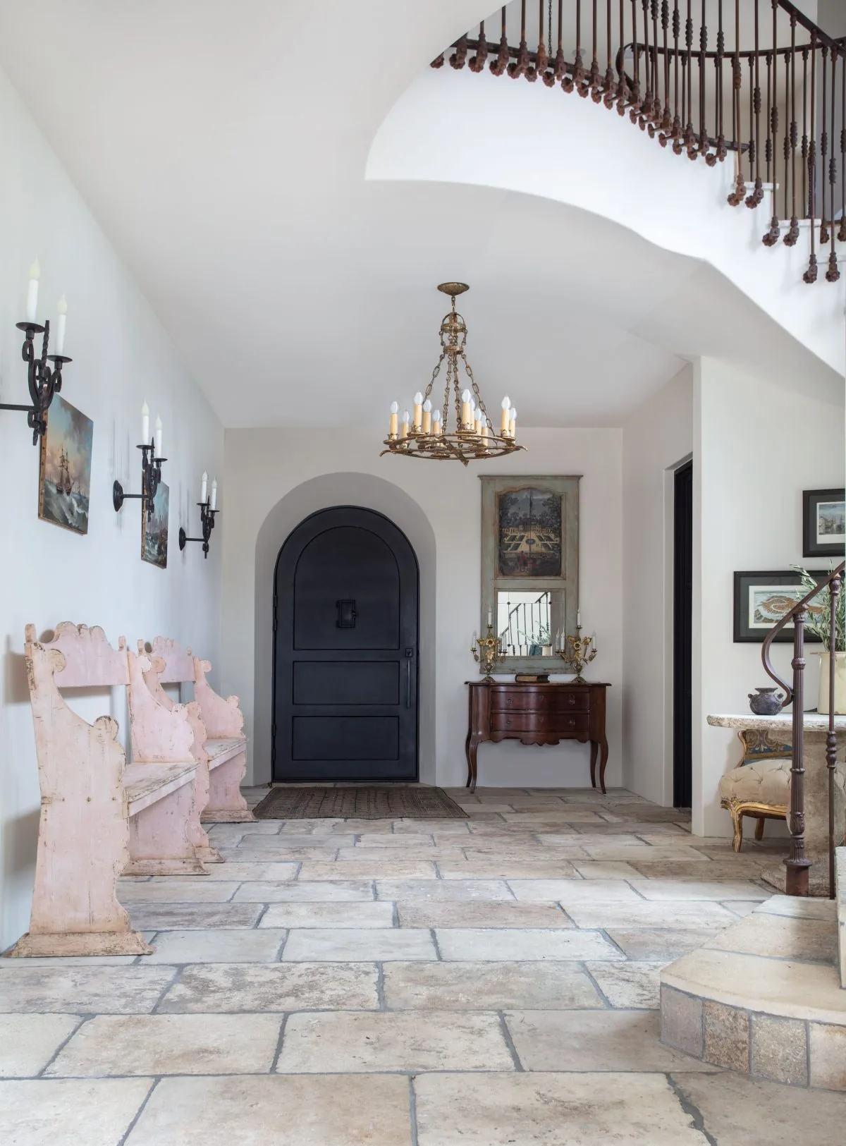 Entry hallway featuring stone tile floors, an arched black front door, wood benches, and a brass chandelier beneath a curved staircase.