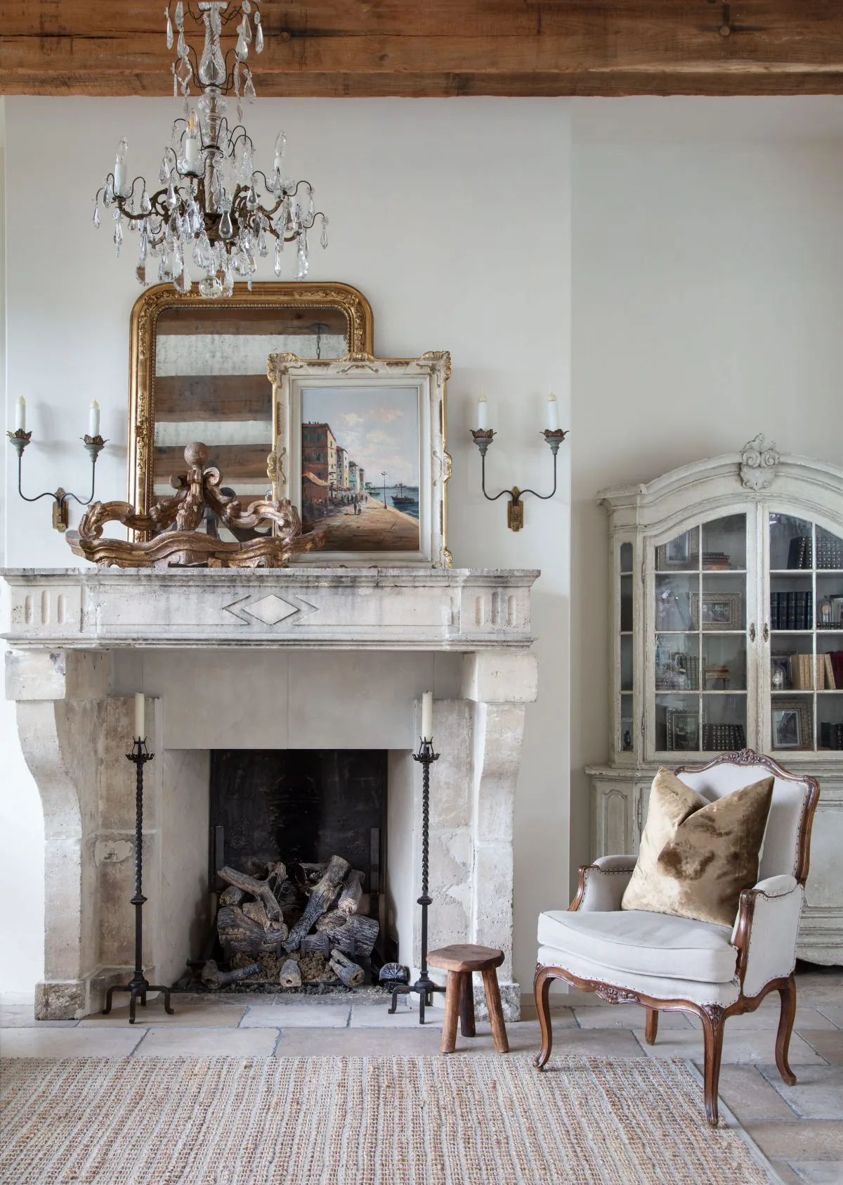 Old-World French estate features a stone fireplace, antique sconces, chandelier, and vintage armchair.