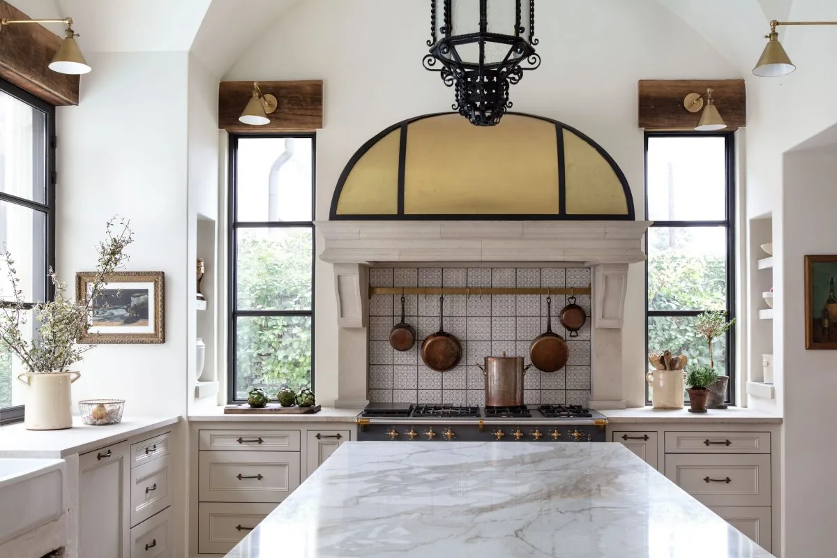 Kitchen in an Old-World French Estate featuring a marble island, white cabinets, and a vintage stove beneath a stone mantel style hood with an arch cover.