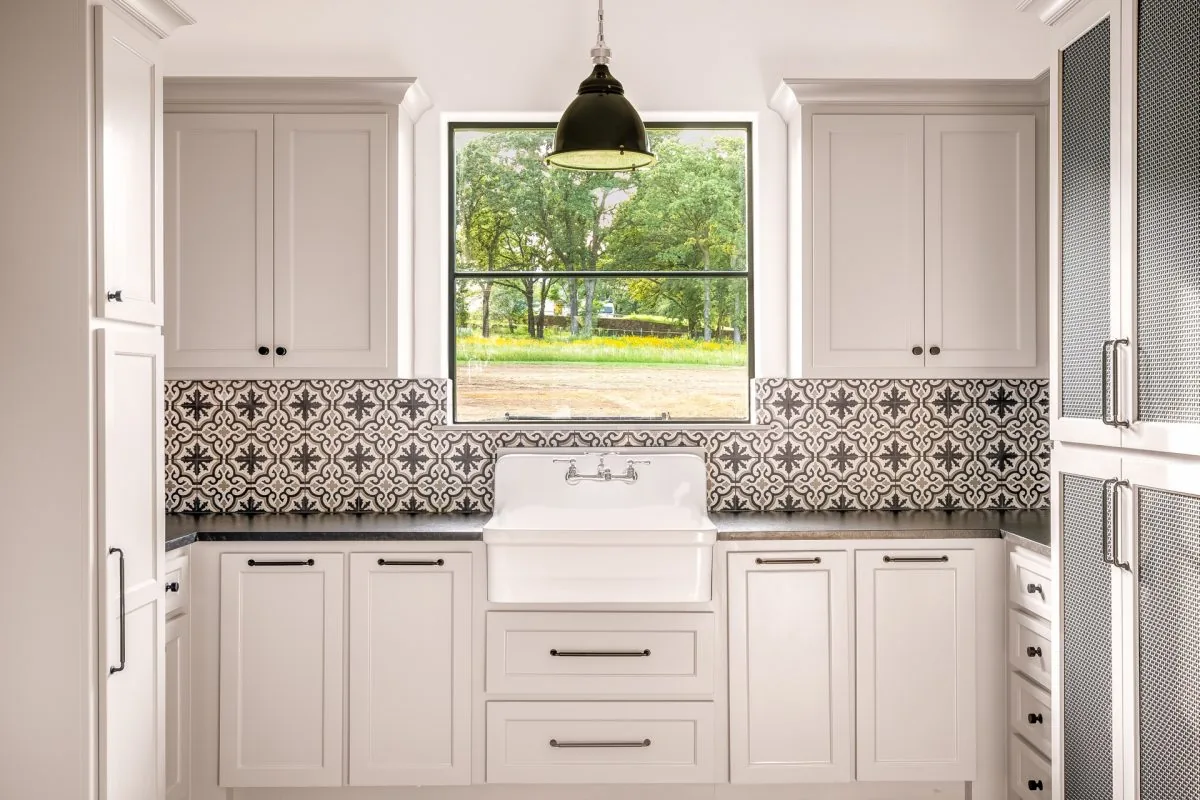 Laundry room features white cabinetry, patterned tile backsplash, a farmhouse sink beneath a large window, and a black pendant light.
