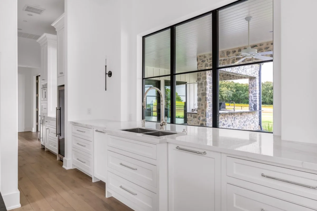 The kitchen features white cabinetry and marble countertops with large black window frames overlooking a stone-clad outdoor area.