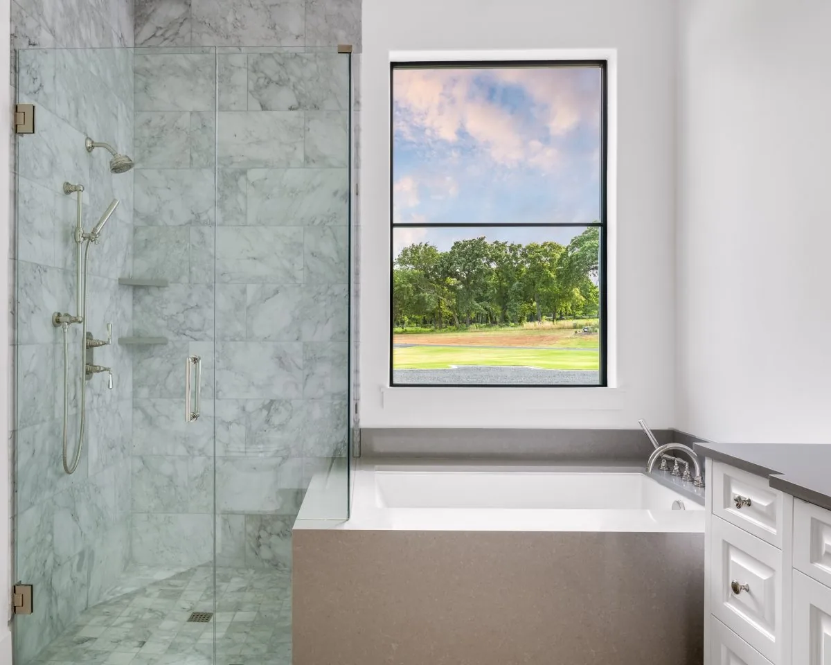 This bathroom includes a marble tile shower, a built-in bathtub, and a large window with black frames providing an outdoor view.