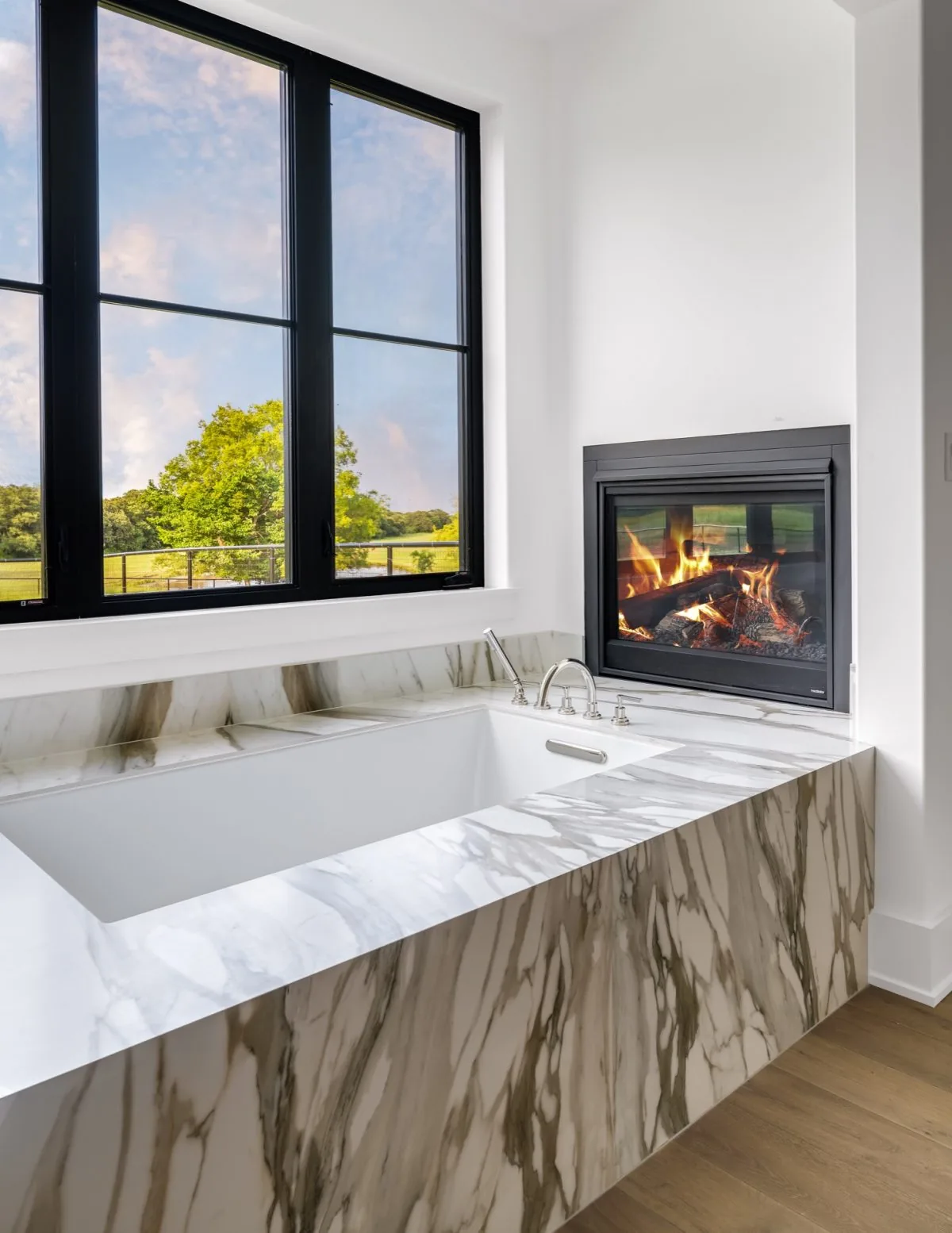 A marble-surrounded bathtub sits adjacent to a fireplace under a large black-framed window in this bathroom.