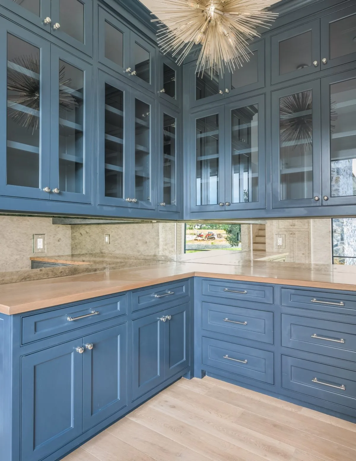 This room corner showcases blue paneled cabinetry with glass doors and a wood countertop beneath a sculptural chandelier.