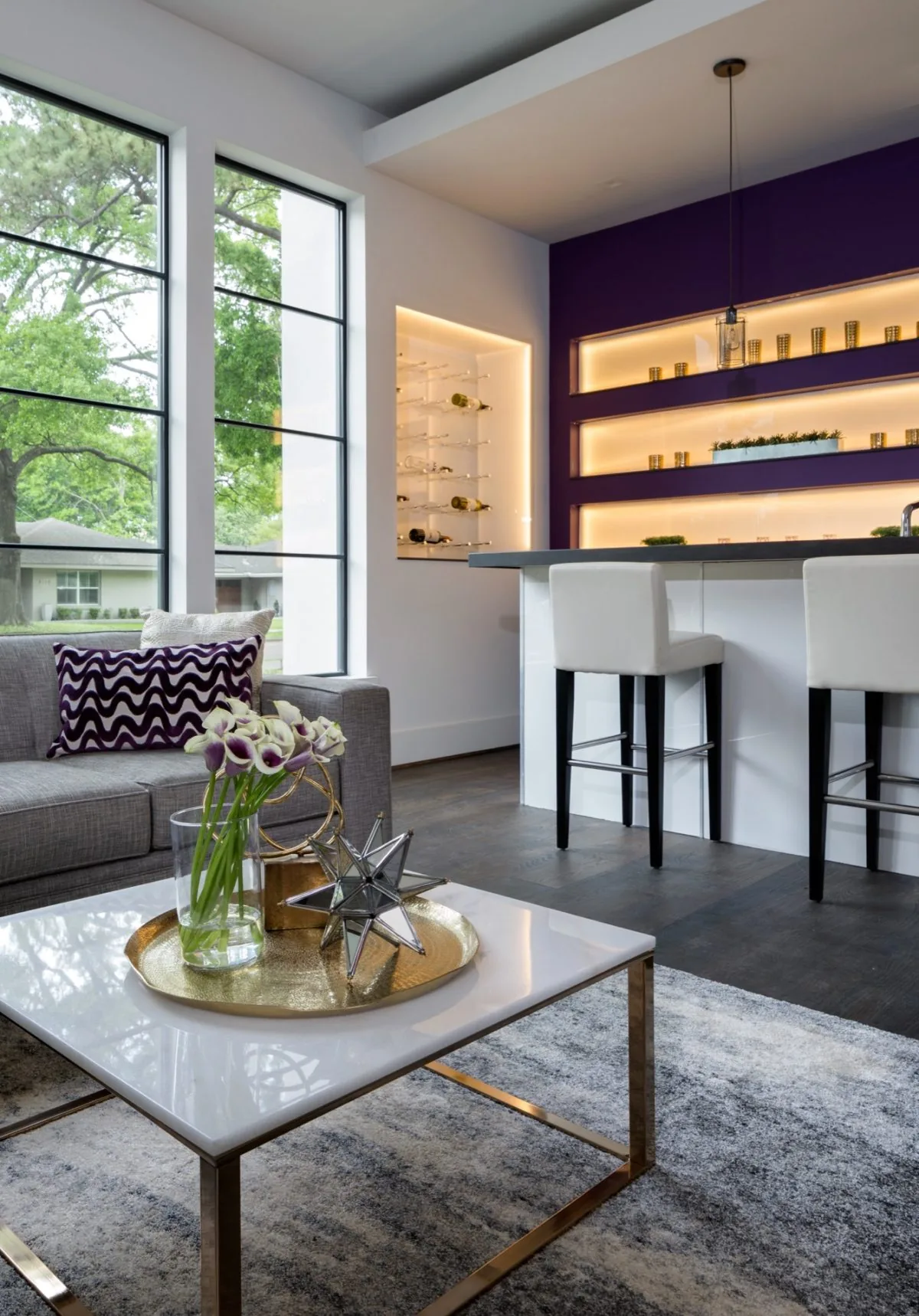 Living space features large vertical windows, a gray sofa, and a kitchen bar with backlit shelves and modern seating.