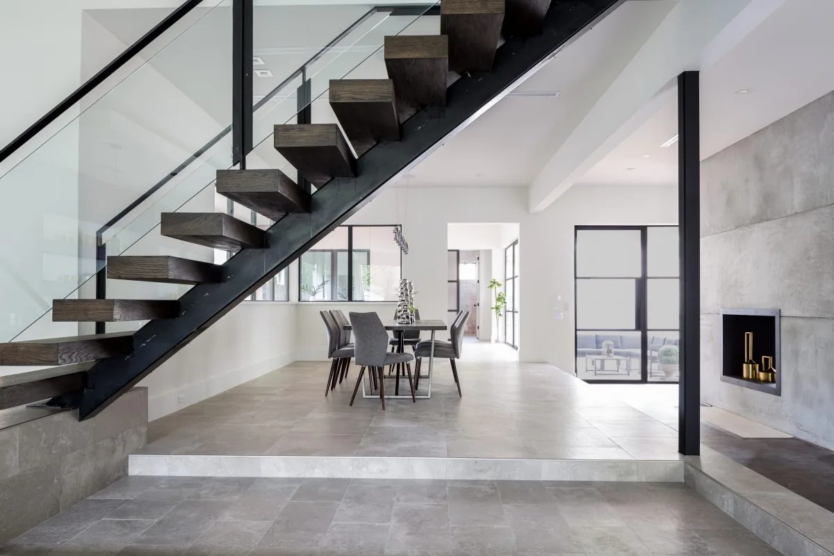 Interior view showing a floating dark wood staircase with glass railing next to a dining area with gray chairs.