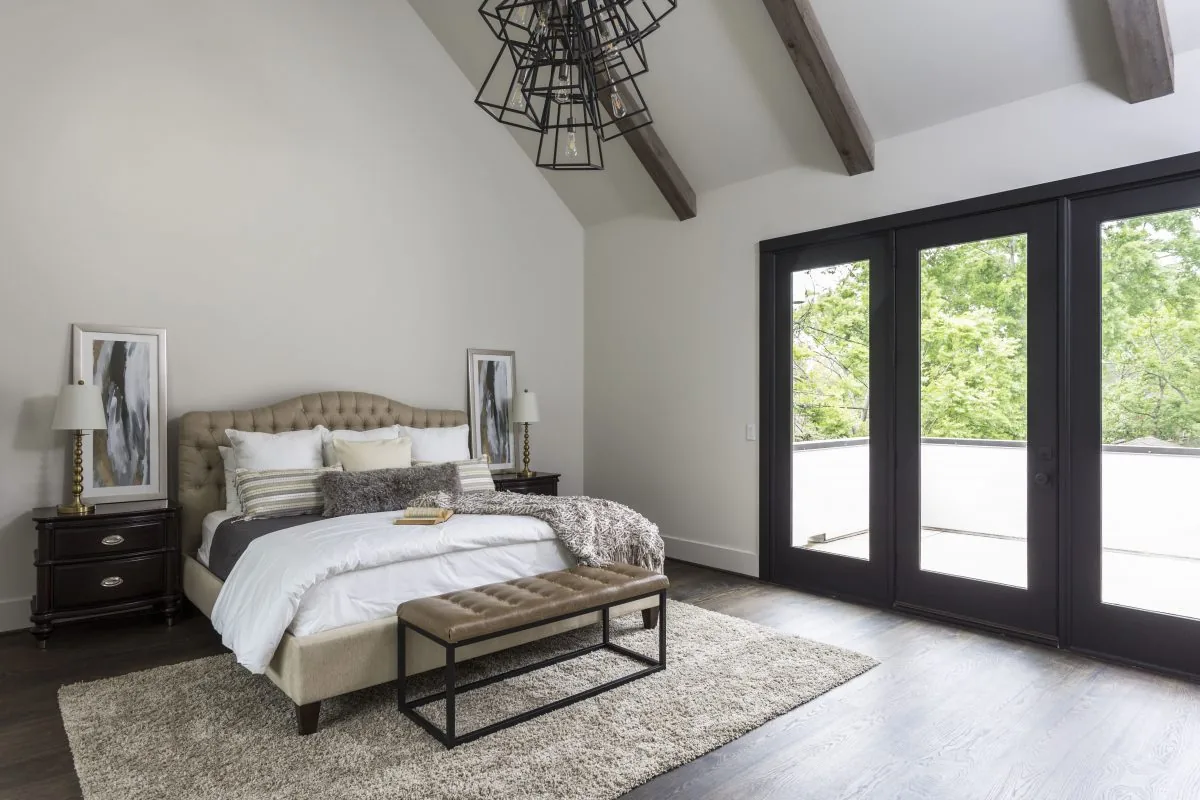 This bedroom features a tufted bed, bench seating, dark wood floors, exposed ceiling beams, and black-framed glass doors to the outside.
