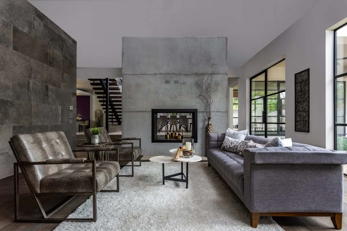 The living area features a concrete double-sided fireplace, gray upholstered sofa, leather chairs, and large black-framed windows.