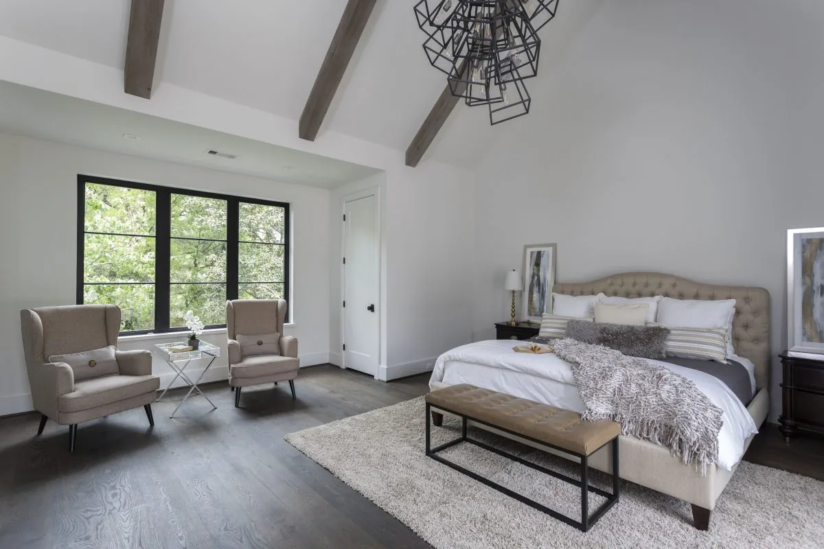 Modern bedroom featuring vaulted ceiling with exposed beams, black-framed windows, tufted headboard bed, and beige armchairs.