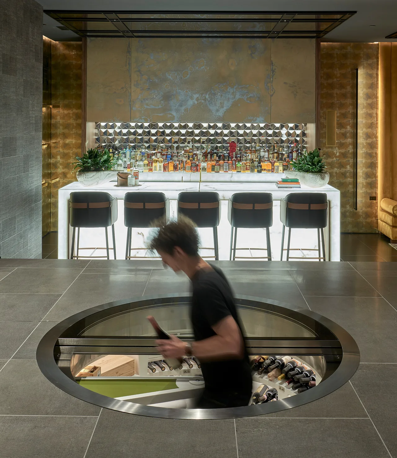 A circular glass floor window reveals a wine cellar beneath, facing a backlit marble bar with geometric mirrored backsplash.