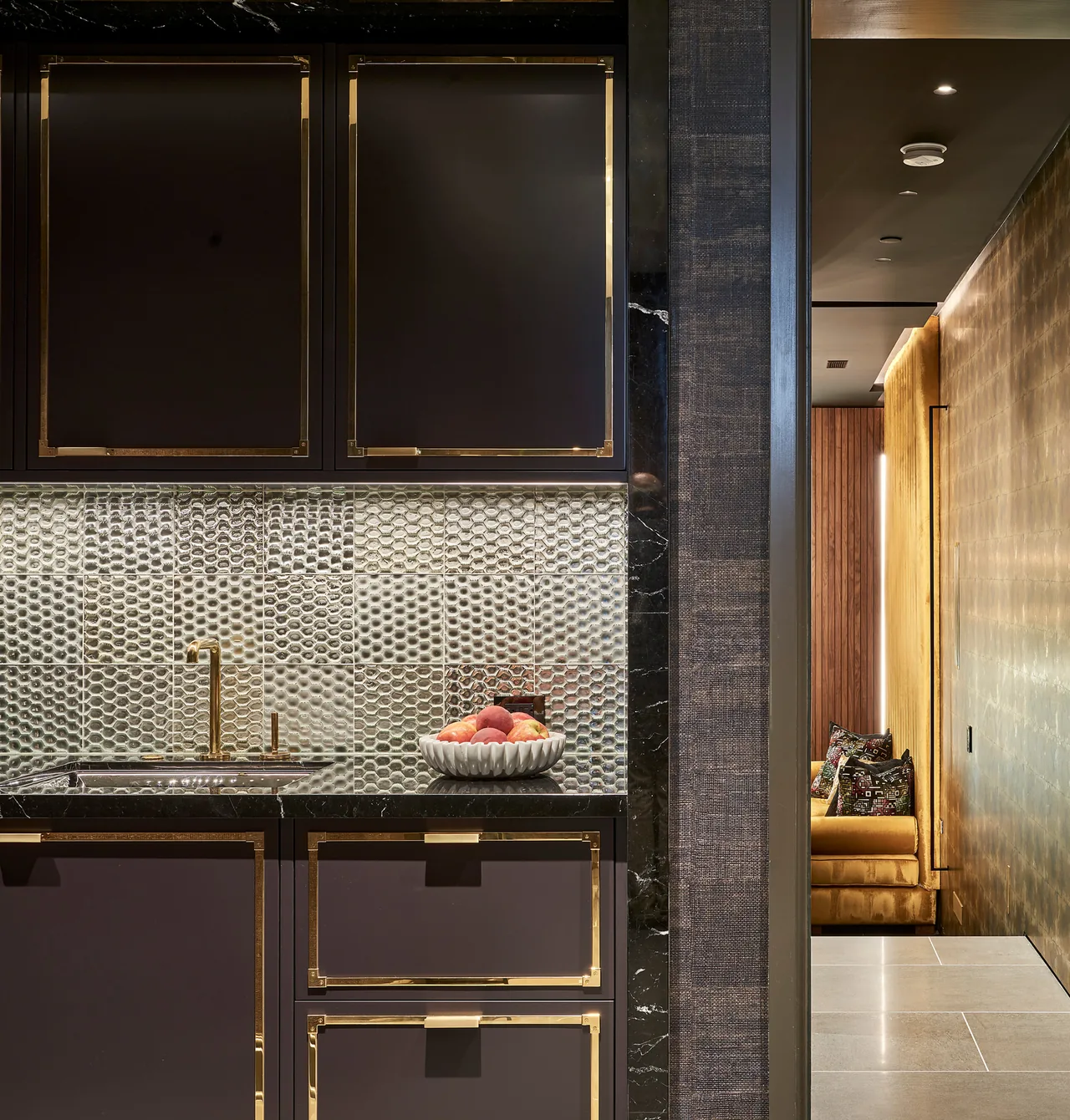 Modern kitchen area showing black cabinetry with gold trim and a textured metallic tile backsplash with integrated sink and faucet.