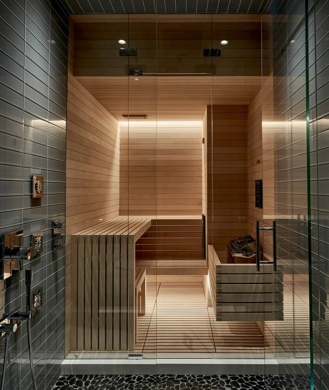 Modern sauna with light wood paneling and benches enclosed by glass door next to a dark tile shower space.
