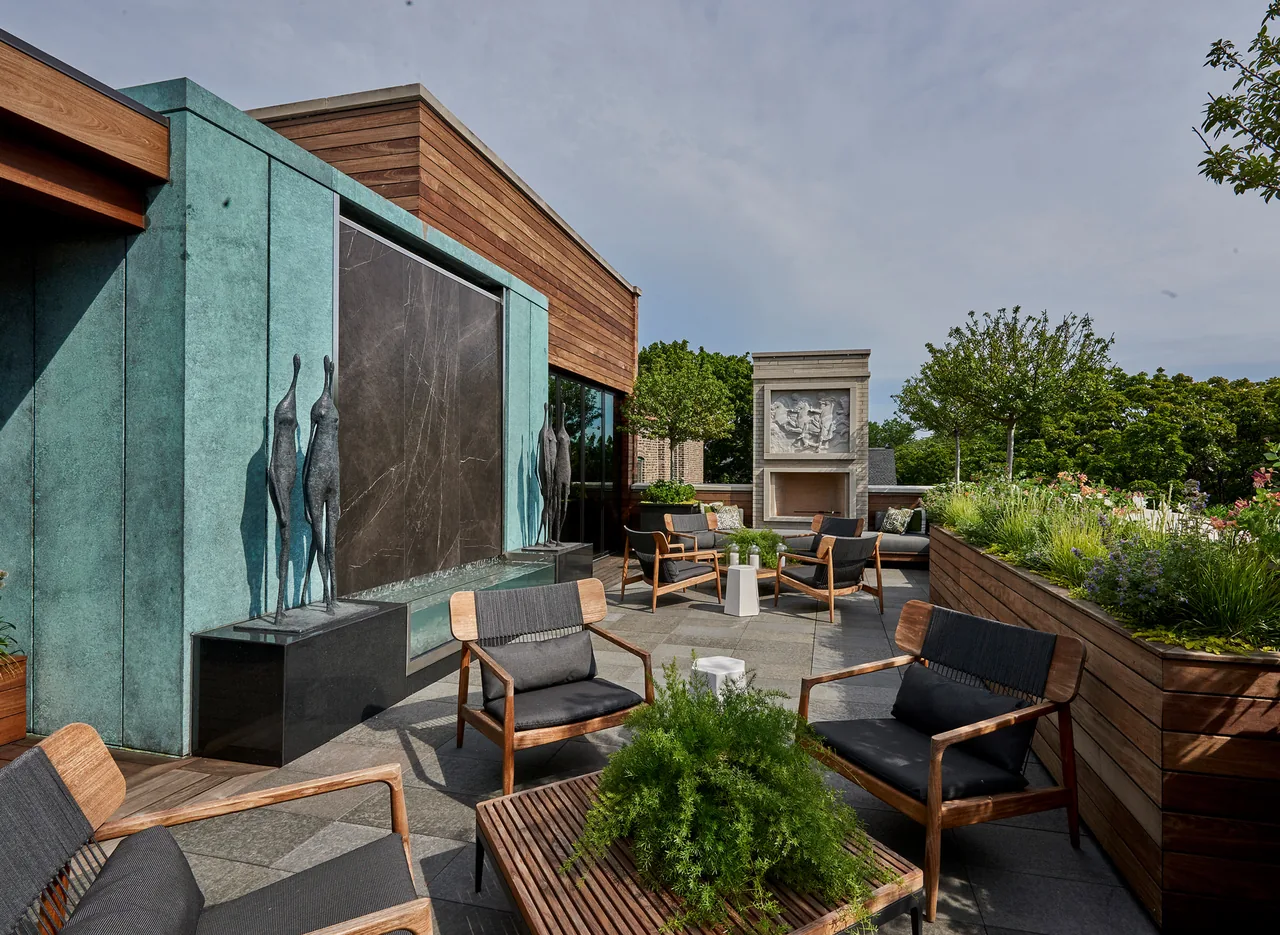 Outdoor living area features wood lounge chairs, a decorative water wall, and a stone fireplace with sculptural relief.