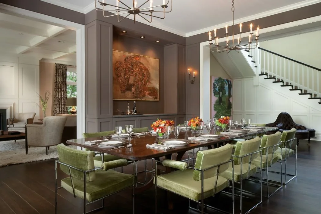 A large dining room features a wooden table with green velvet chairs and a modern chandelier in a home with an open entry to a living room and staircase.