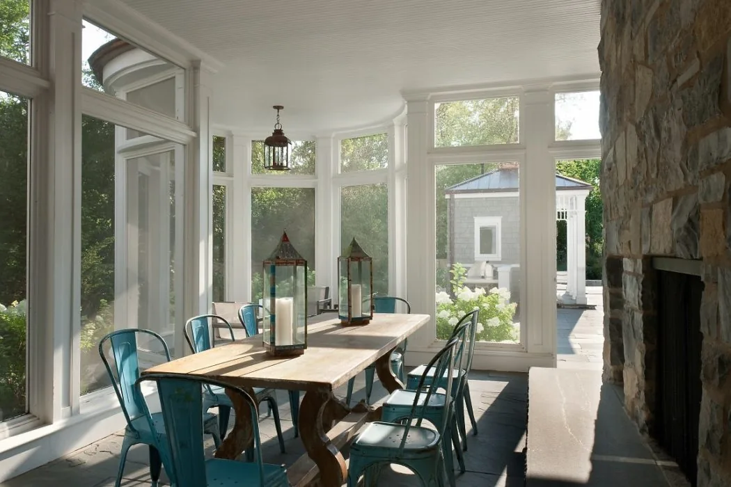 Sunlit dining area in a sunroom with a wooden table, blue metal chairs, and a stone fireplace wall.