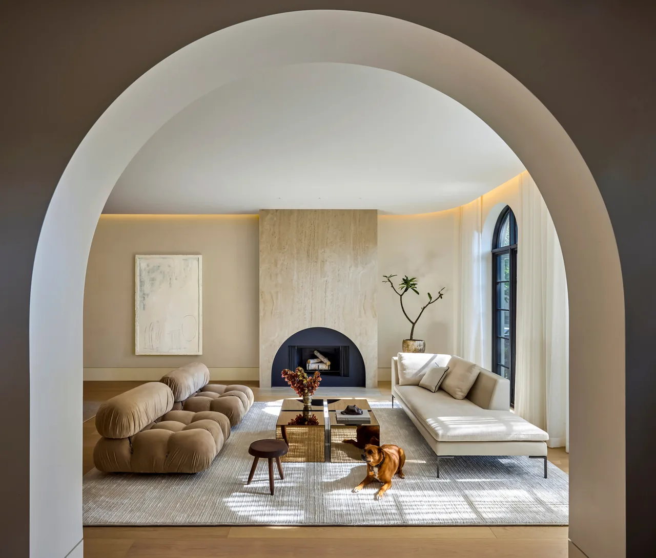 Contemporary living room with arched entry, cushioned tan sofa, white chaise, large light stone fireplace, and floor-to-ceiling arched window.