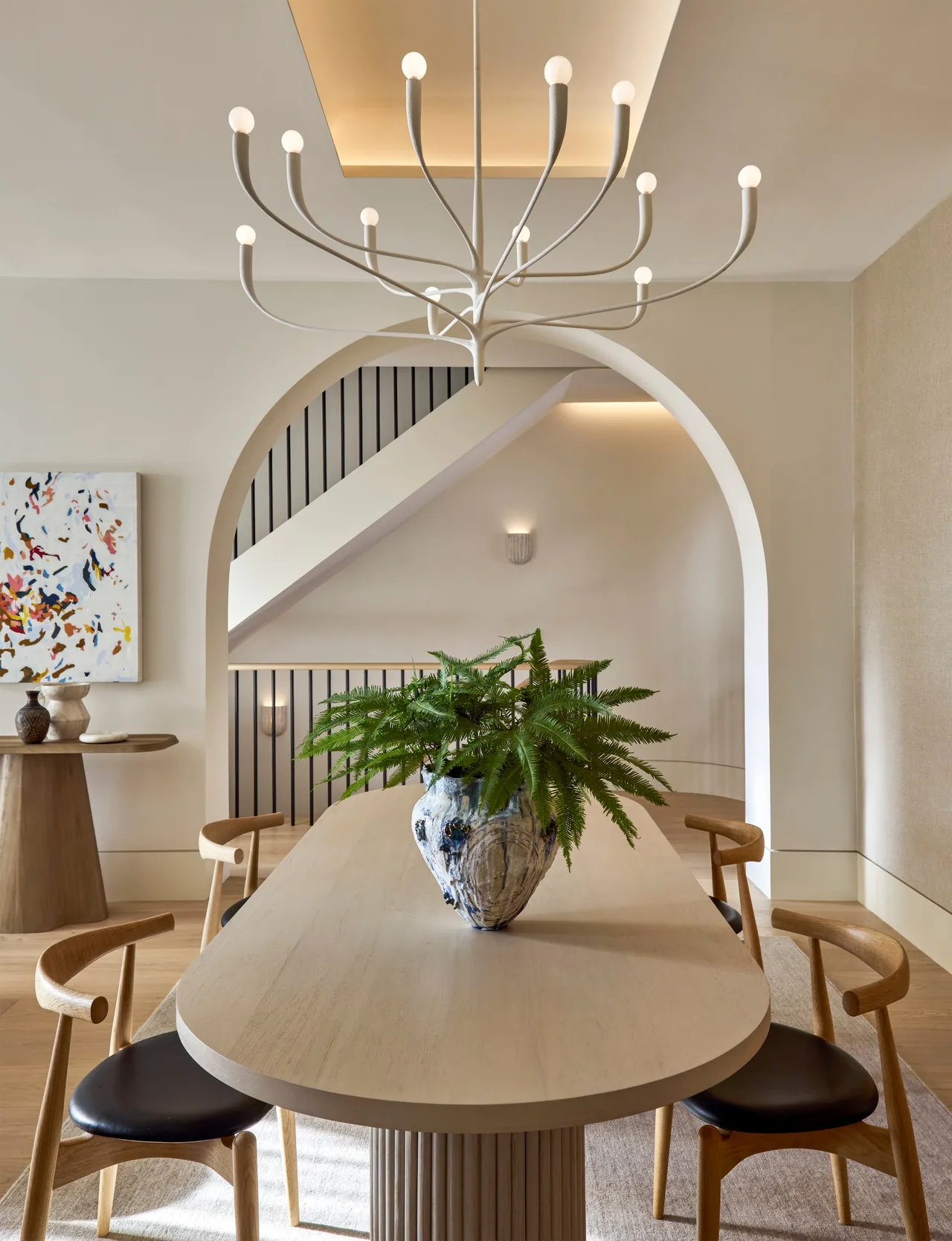Dining area features a light wood table with black chairs under a modern multi-arm chandelier framed by an arch overlooking a staircase.