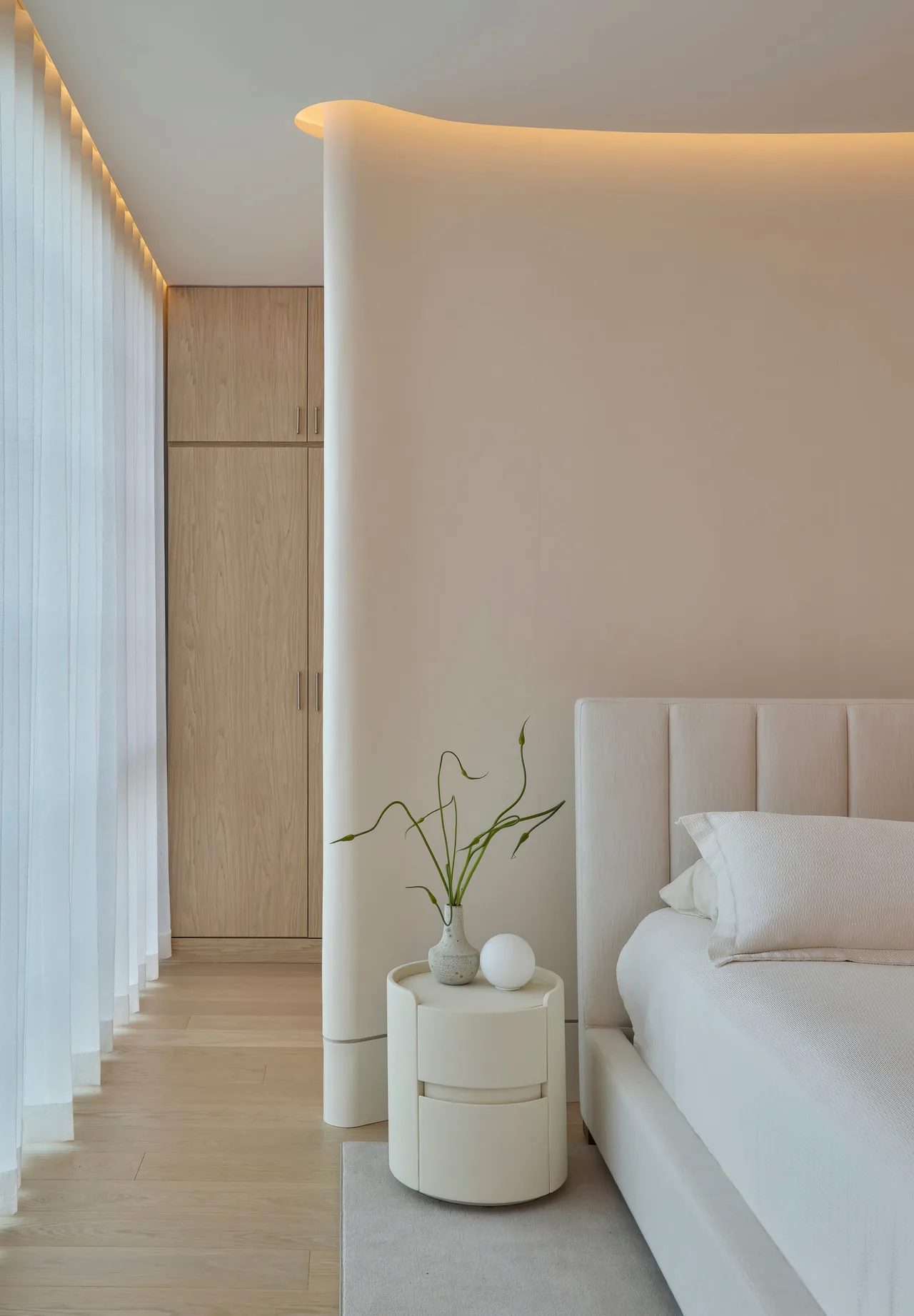 Bedroom with a tufted upholstered bed, a curved wall featuring indirect cove lighting, and a round white nightstand with a vase.