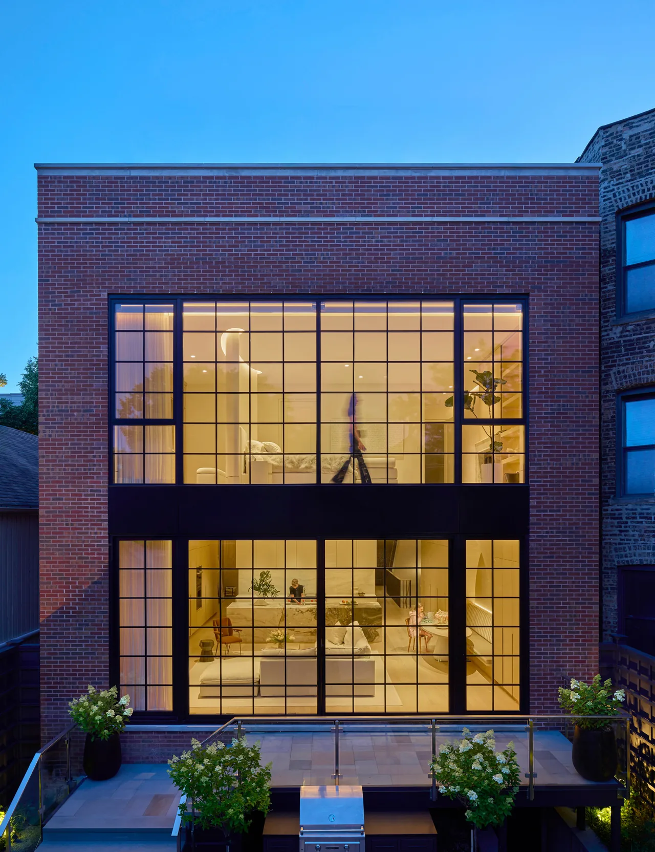Two-story brick facade with expansive black steel windows reveals illuminated living and bedroom areas.