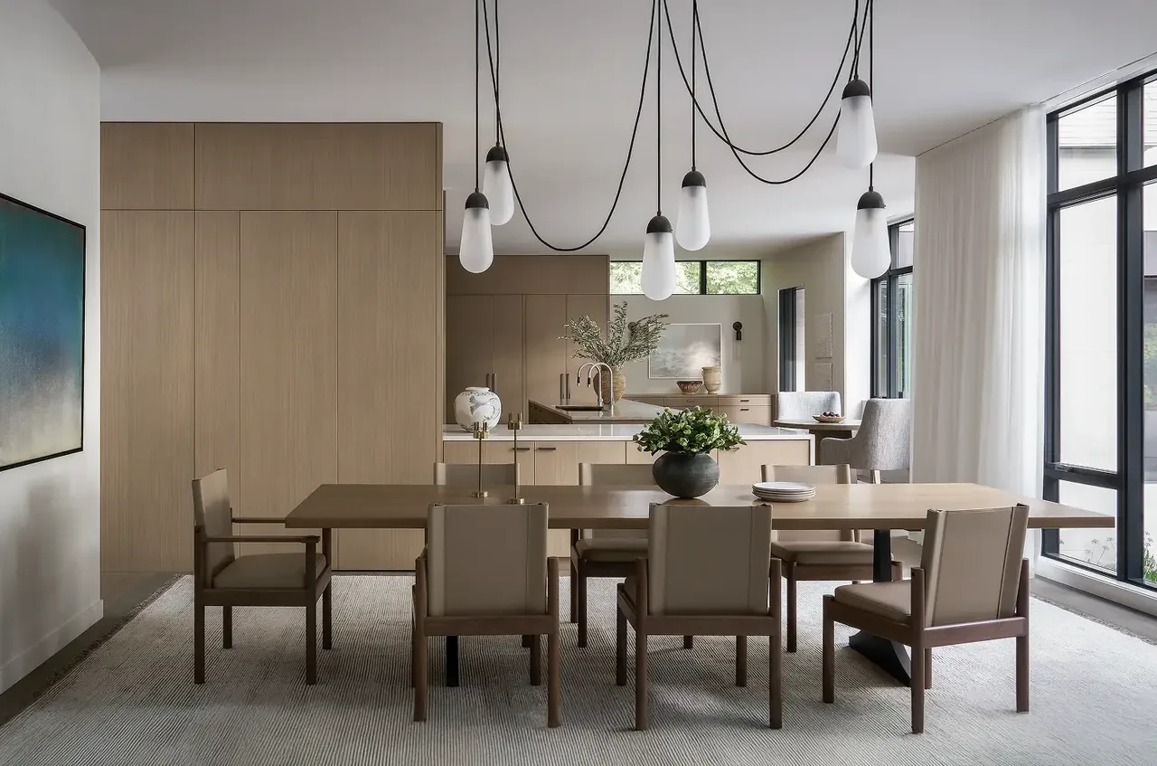 Dining room with beige wood table and chairs, minimalist pendant lights, and large black-framed windows next to an open kitchen with light wood cabinetry.