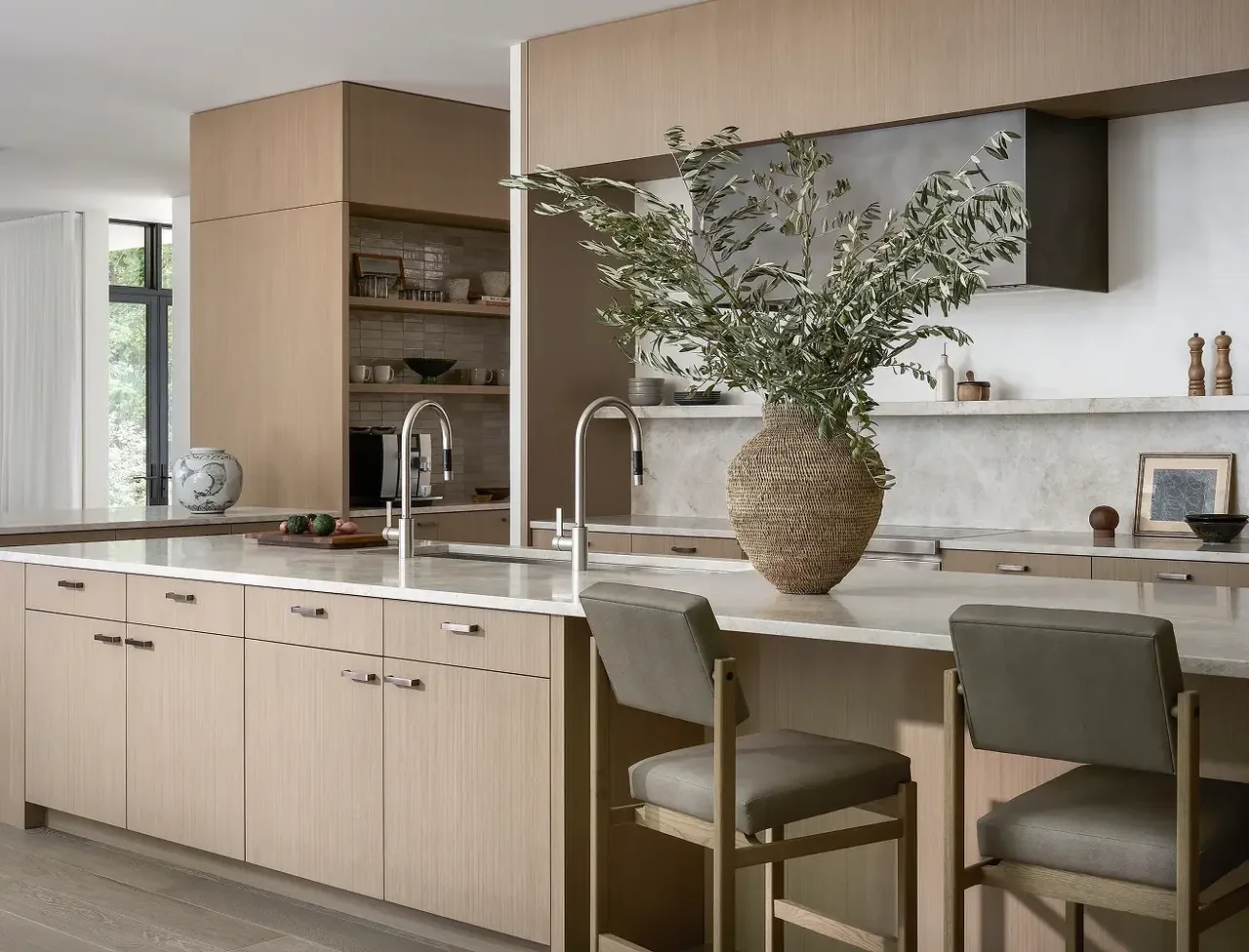 The kitchen of Ravine Reimagined features a large island with two sinks and light wood cabinets, accented by a natural woven vase with greenery.