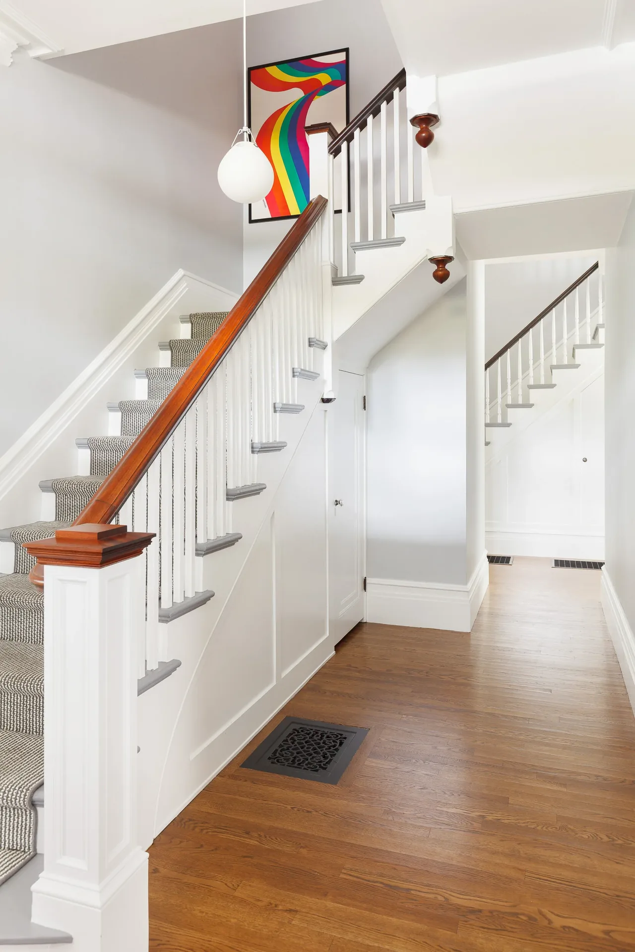 A bright hallway features a wood-handled staircase with white spindles, gray-trimmed steps, and hardwood floors.