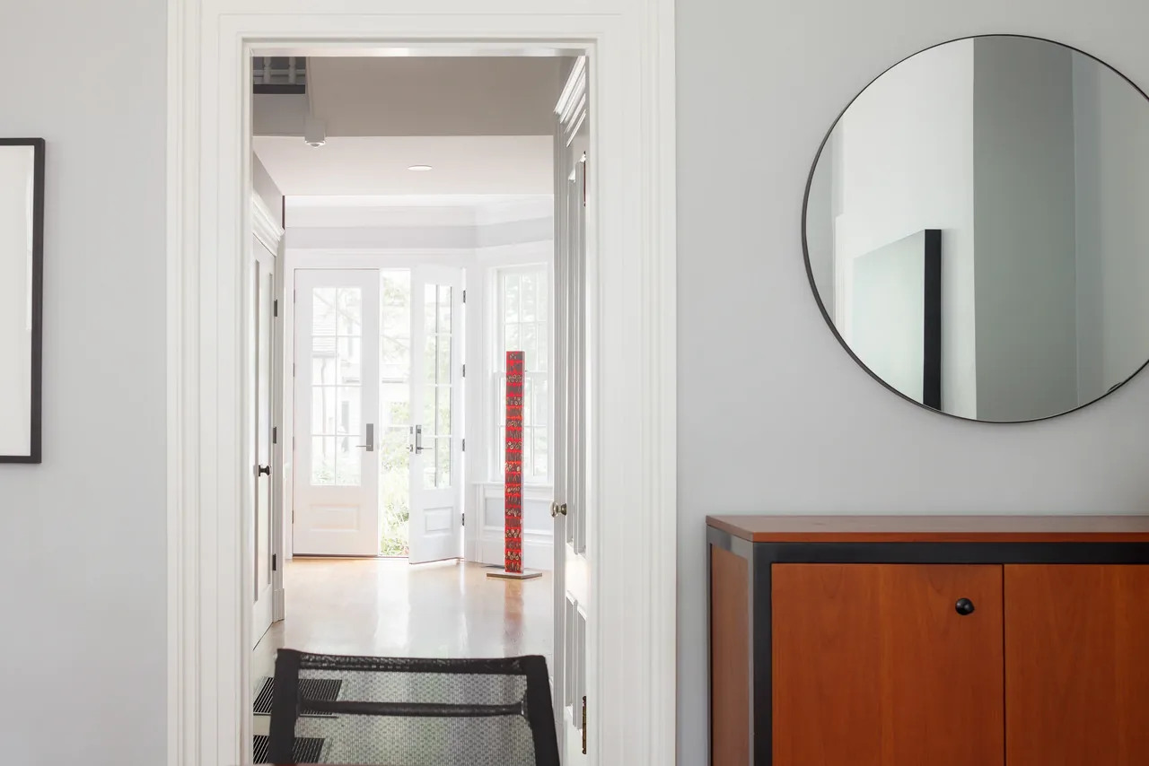 Interior view through white trim doorway into a sunlit room with wood flooring and a red vertical sculpture near a glass door.