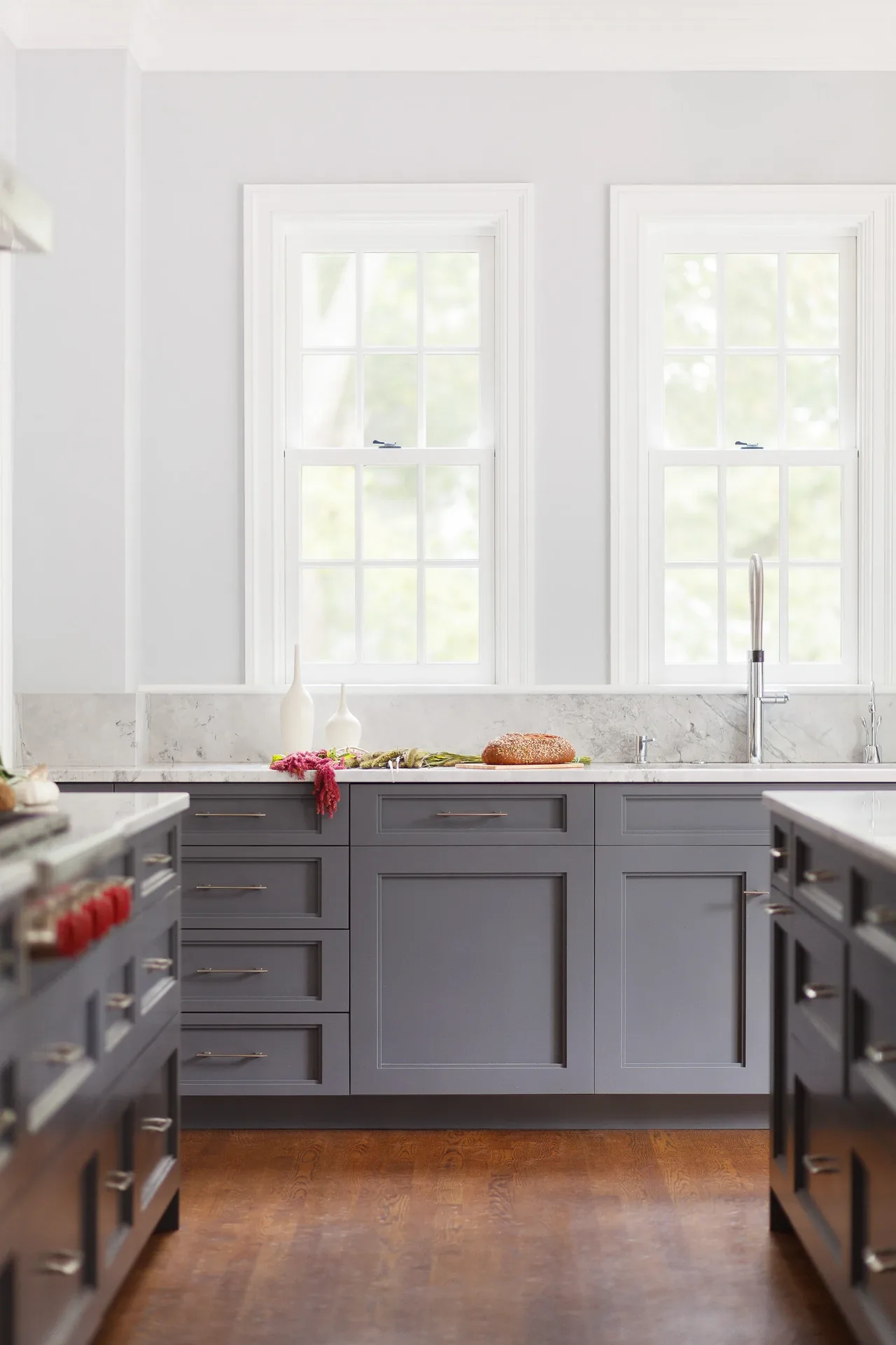 Kitchen featuring gray cabinetry and marble countertops beneath two white-framed windows.