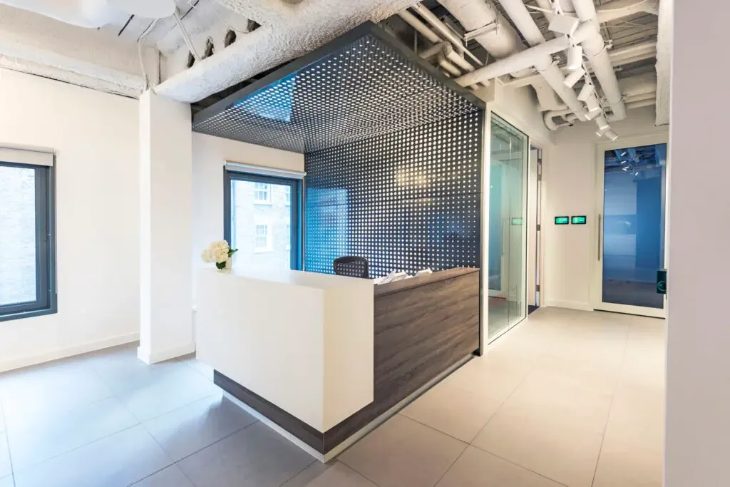 Reception area features a wood and white desk against a perforated black metal wall with large windows and exposed ceiling piping.